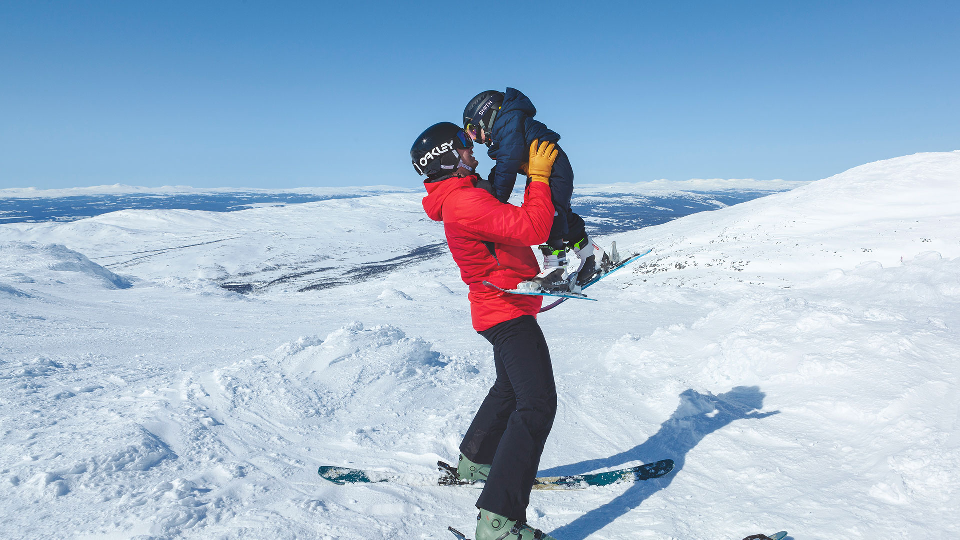 Person i röd jacka lyfter barn med hjälm och skidor på snötäckt fjäll