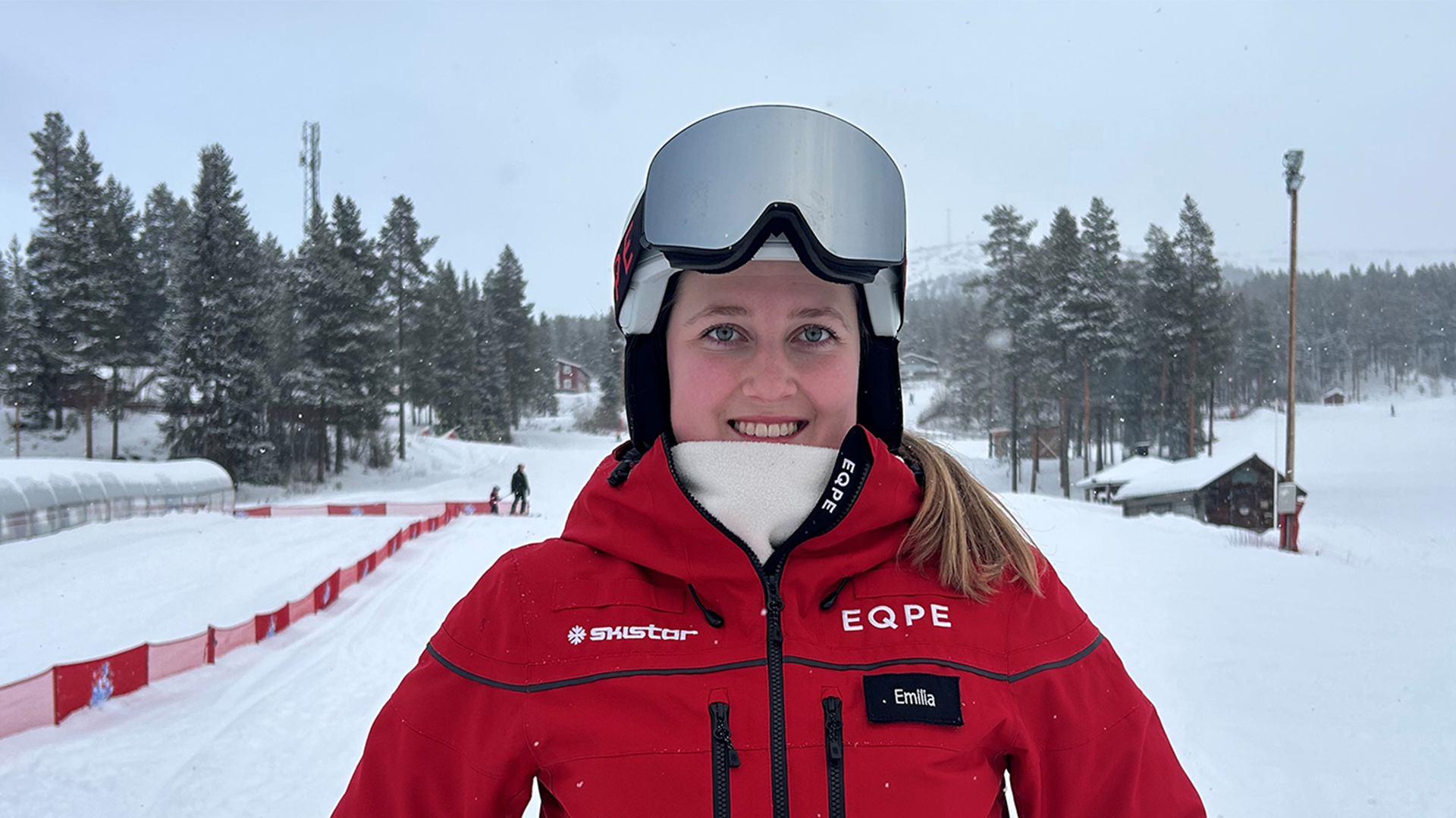 Kvinna i röd skidjacka och skidglasögon står på en snötäckt skidbacke med skog i bakgrunden