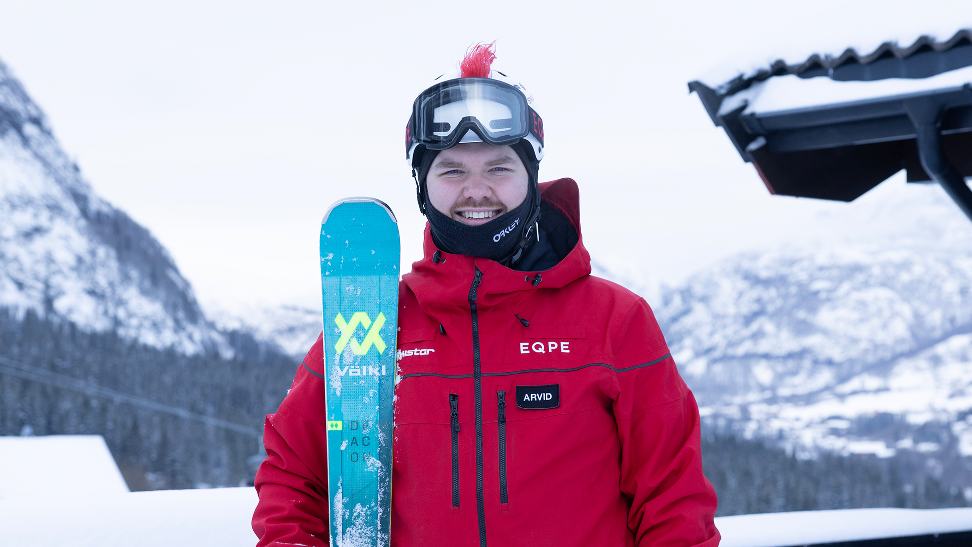 Person i röd skidjacka och skidglasögon håller en Völkl-skida, med snöiga berg i bakgrunden. Perfekt för vintersport och skidåkning