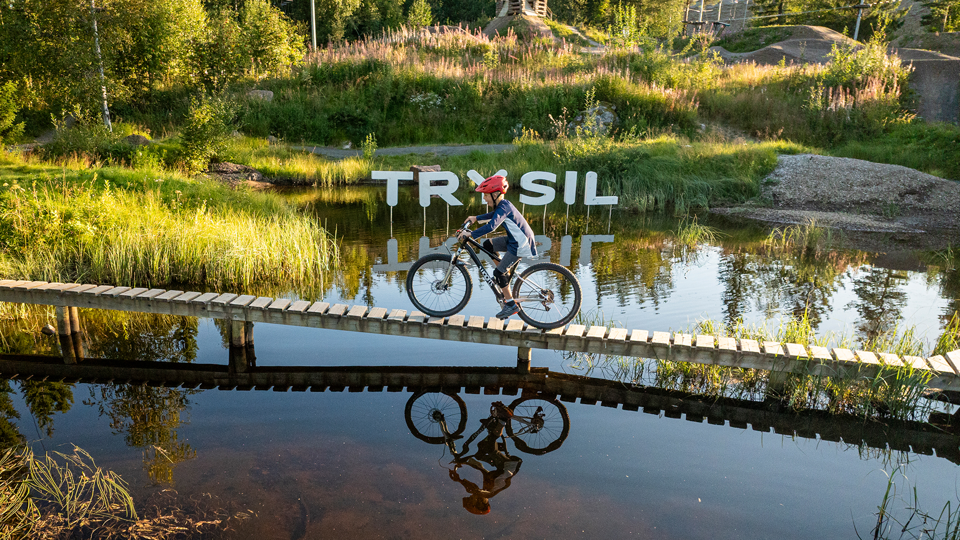 En pojke cyklar på en spång över ett vattendrag och solen lyser.