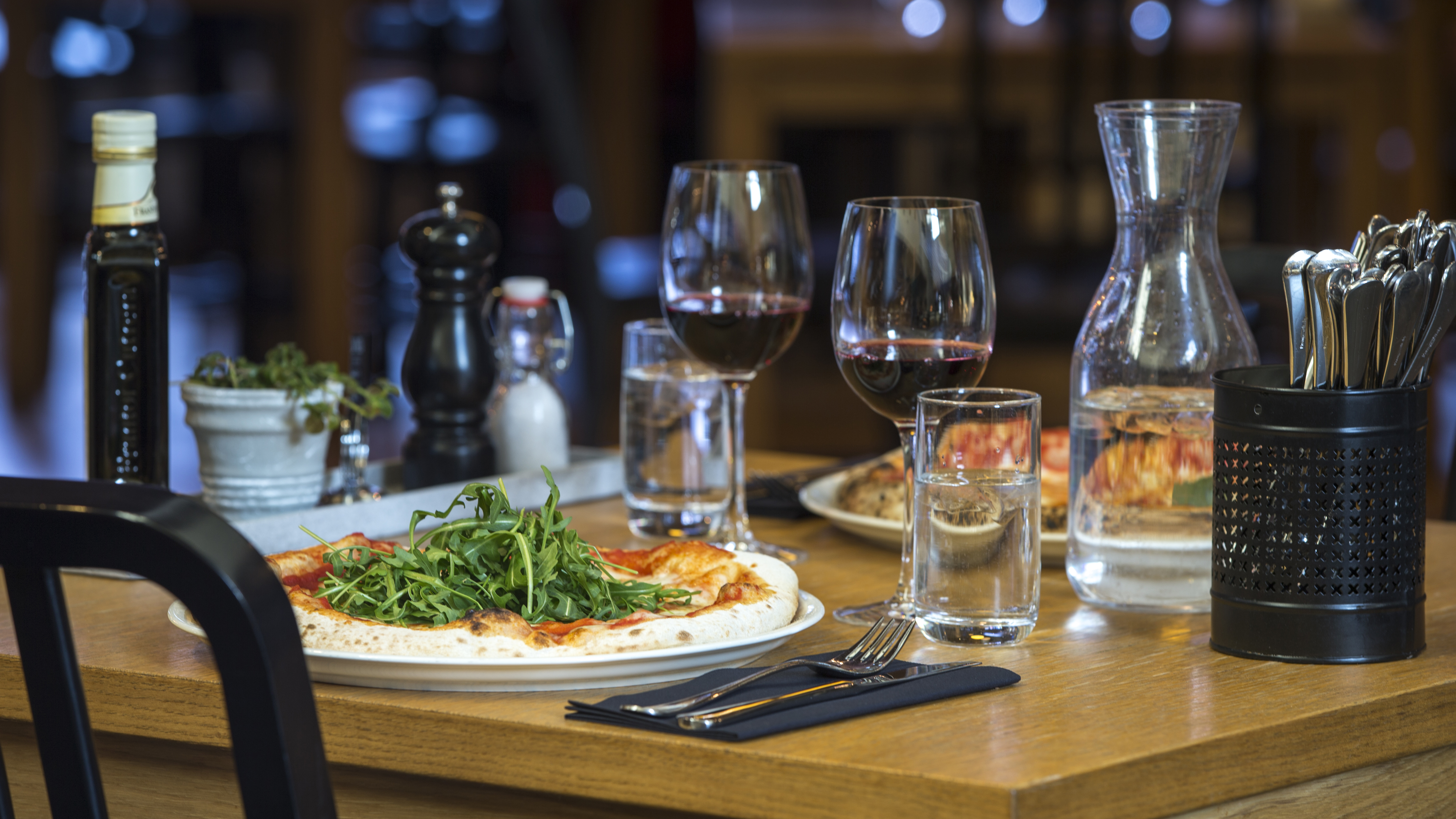 Pizza med ruccola serverad på ett träbord tillsammans med ett glas rödvin, vattenkaraff och bestick