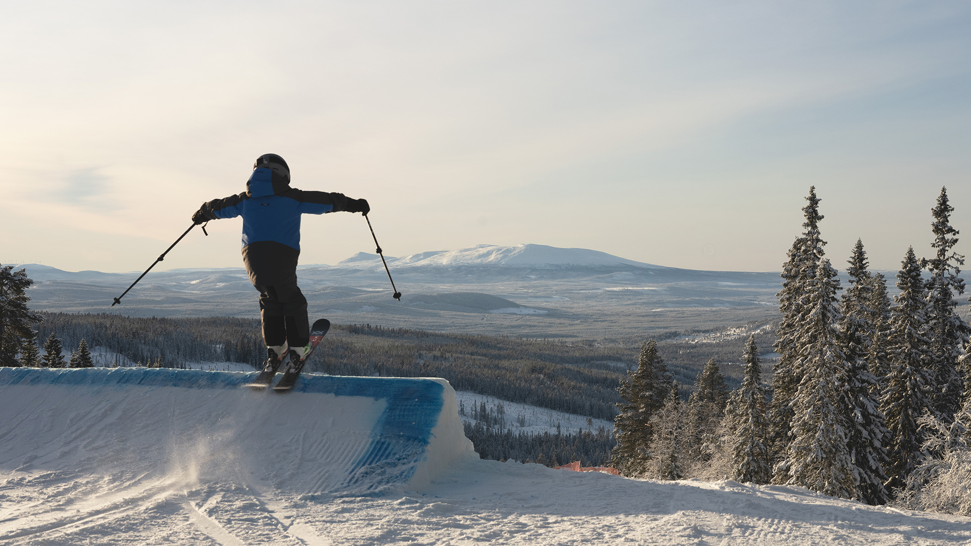 Person åker skidor på ett blått rail i en snötäckt skidbacke med granar och berg i bakgrunden under en klar himmel