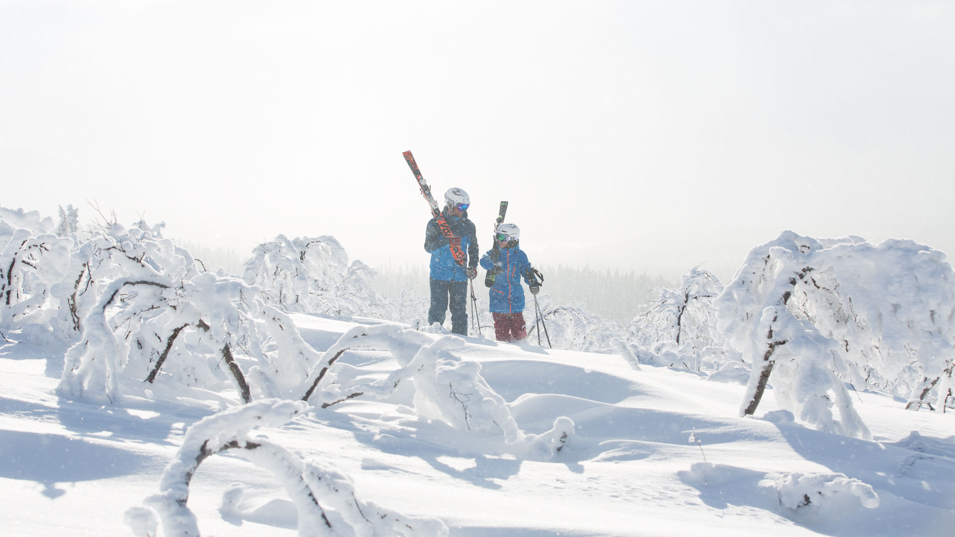 Två personer klädda i vinterkläder står med skidor i en snötäckt skog, omgivna av snötyngda träd och klar himmel