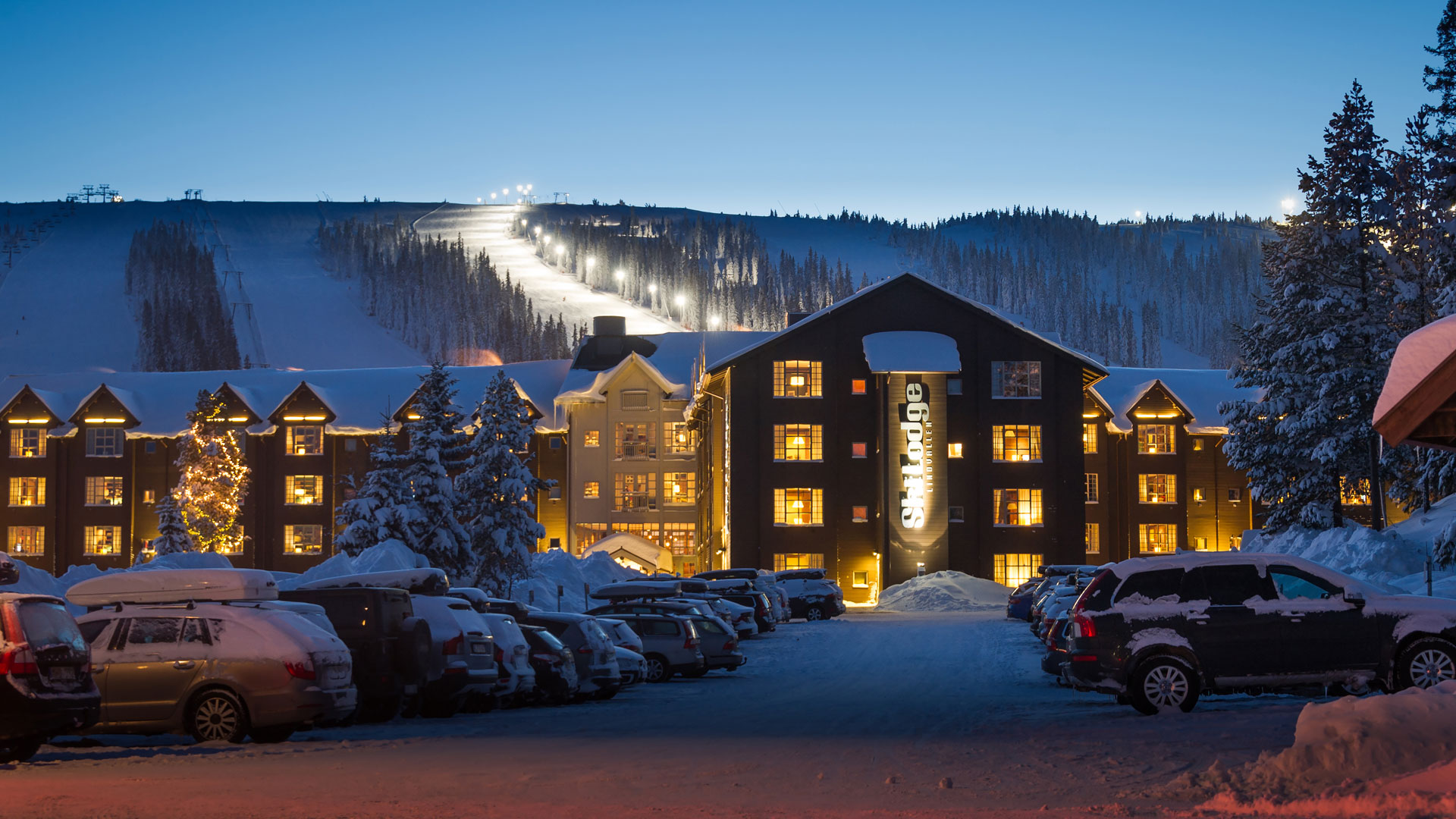 Skihütte und beleuchtete Skipiste in Winterlandschaft, Abendbeleuchtung, Parkplatz mit Autos, die mit Schnee bedeckt sind