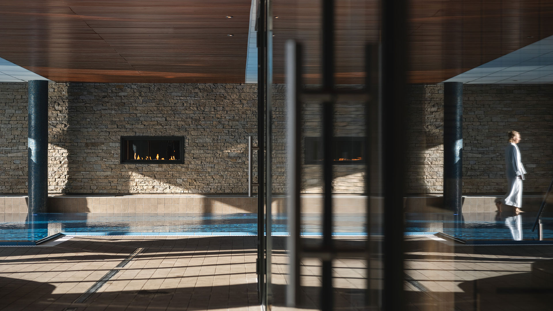 Lugn spa-miljö med en kvinna i badrock som går vid en inomhuspool och en vägg med öppen spis