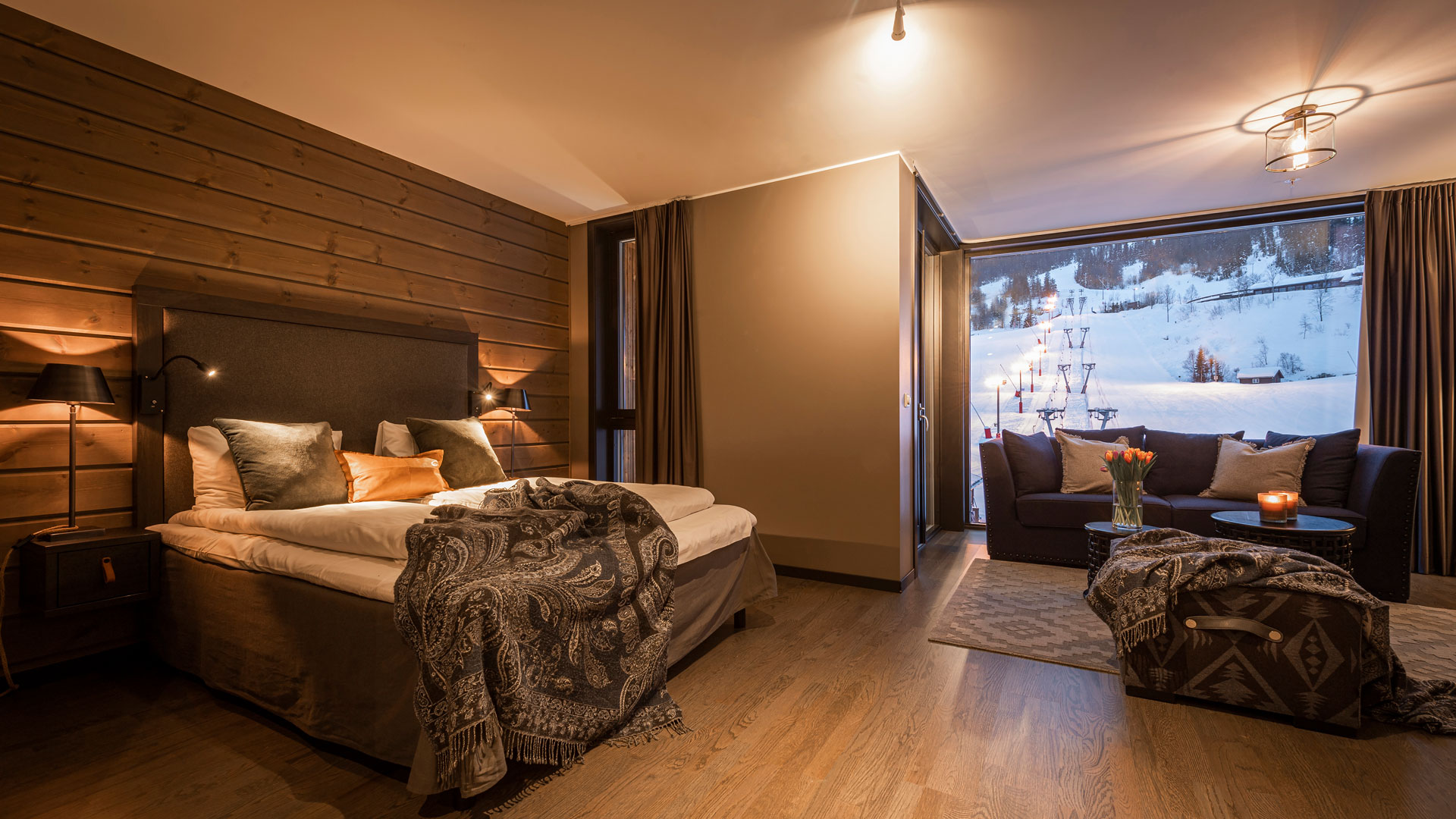 Modernt hotellrum med utsikt över snöklädda berg. Rummet har en stor säng, mörka träpaneler, soffgrupp och dekorativa filtar