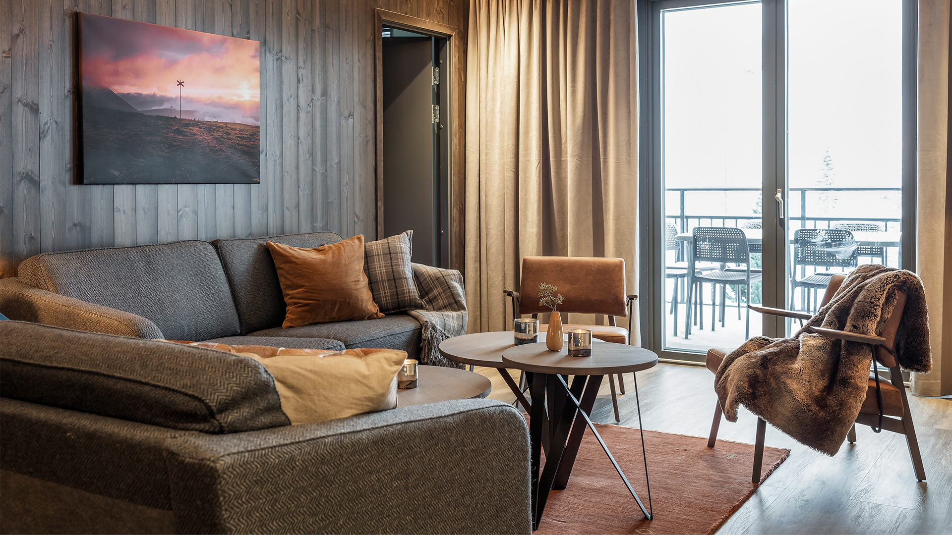 Modernt och mysigt vardagsrum med grå soffa, bruna kuddar, dekorativ tavla, fönster med gardiner och en öppen balkong med utemöbler