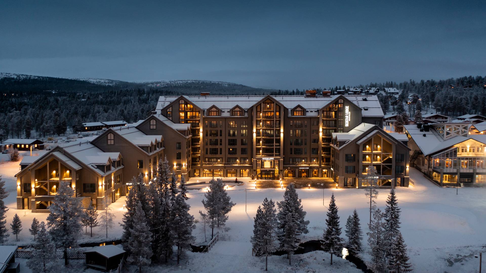 Hotell i vinterlandskap med snötäckt mark och byggnader upplysta i skymningen, omgiven av frostiga träd och berg