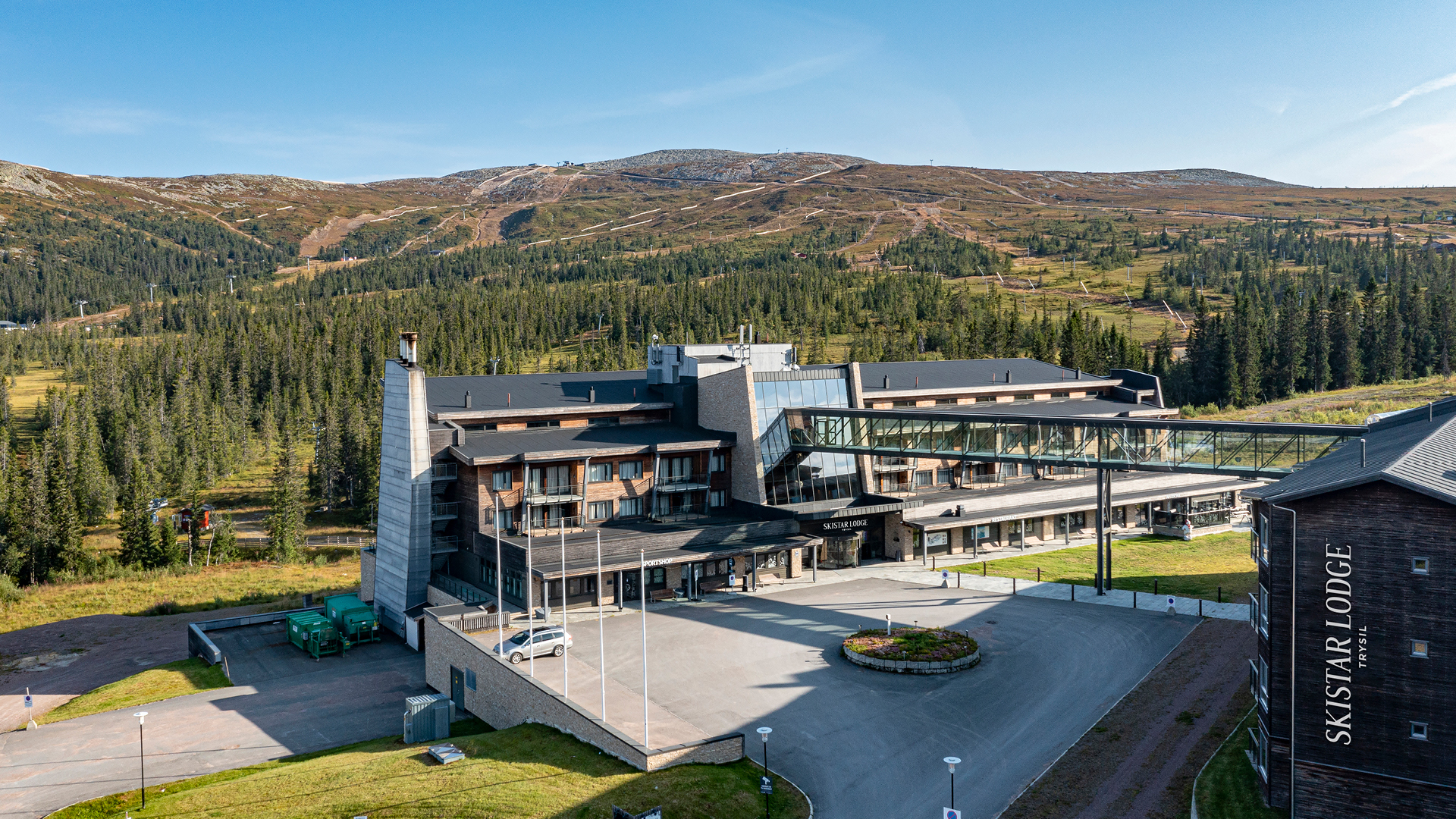 SkiStar Lodge Trysil i fjällmiljö med omgivande skogar och berg. Modernt hotellkomplex med gångbro och parkering i förgrunden