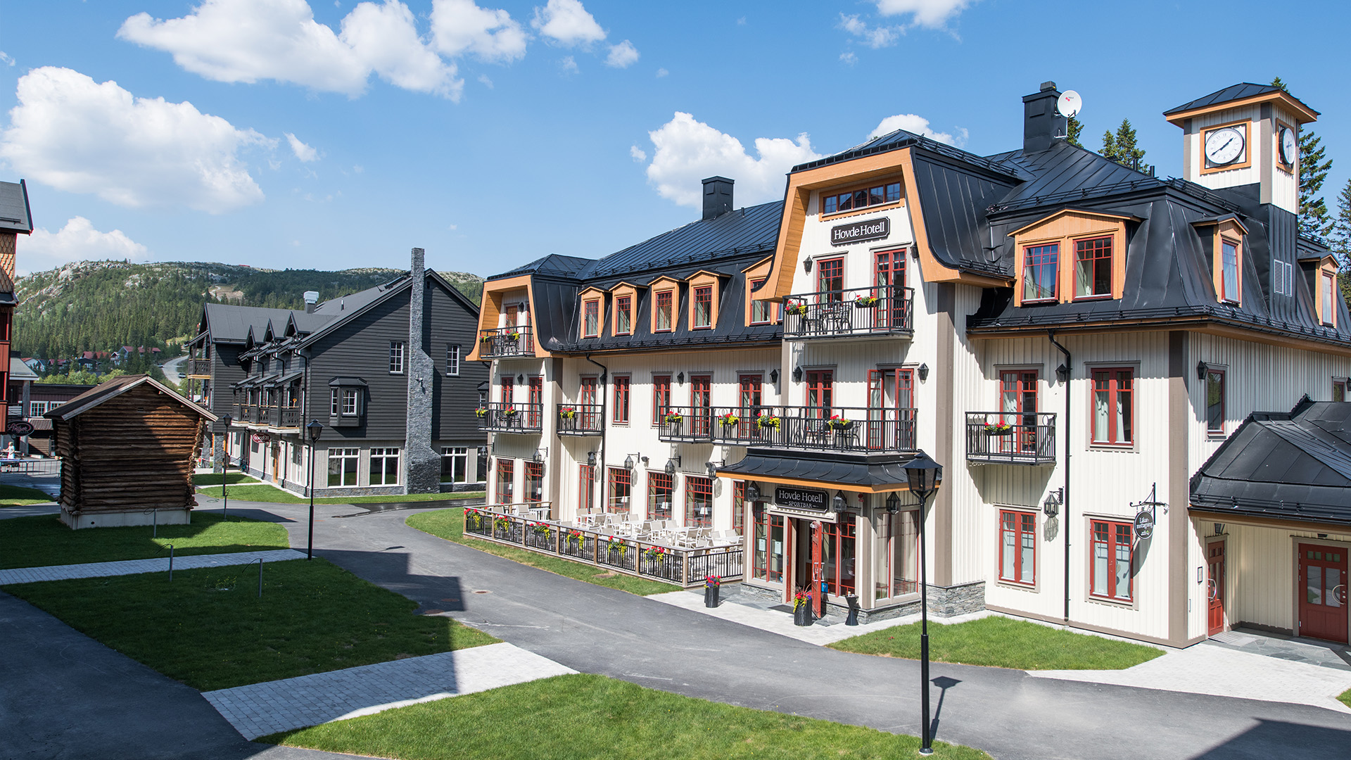 Vy över ett charmigt hotellkomplex i fjällmiljö, med traditionell arkitektur, gröna gräsmattor och en klarblå himmel i bakgrunden i det natursköna området Vemdalsskalet, Sverige, Hovde hotell