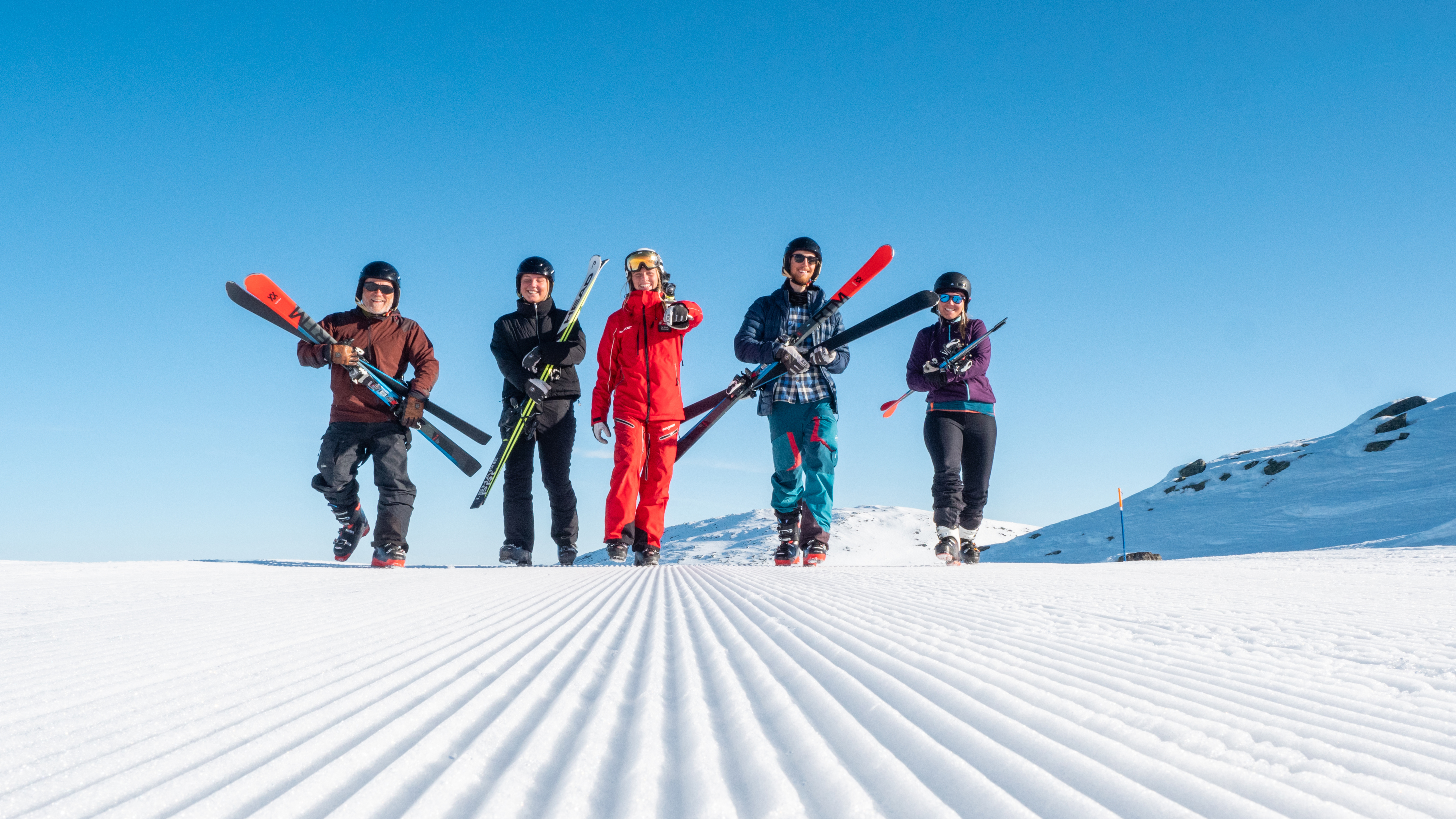 Fem personer i skitøj går på en snedækket pist under en klarblå himmel, mens de bærer deres ski. Perfekt vejr til skiløb i fjeldene