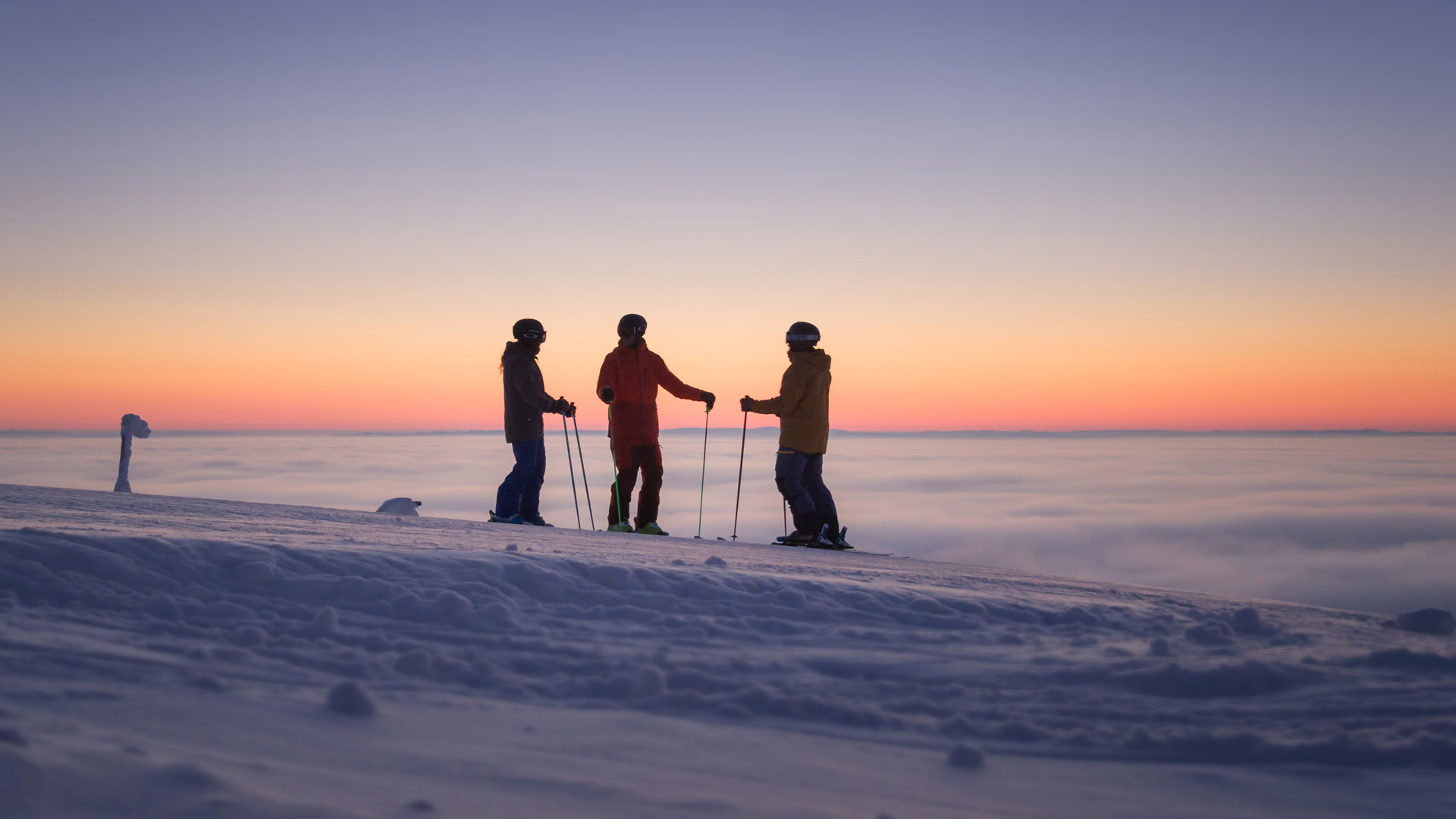 Tre personer i skidkläder står på en snöig bergstopp vid solnedgången, med molntäcket nedanför och himlen i pastellfärger