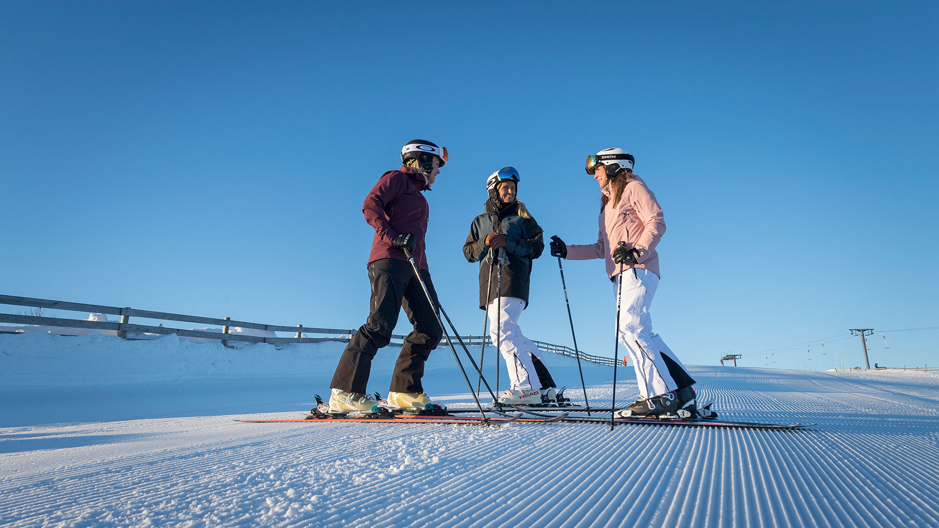 Tre personer åker skidor på en solig dag i en skidbacke med klarblå himmel. De bär hjälmar och skidutrustning