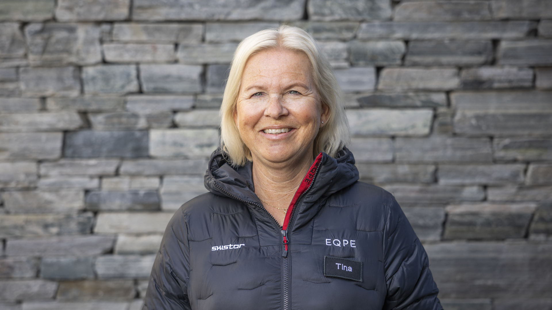 Kvinne i svart jakke med EQPE-logo foran en steinmur