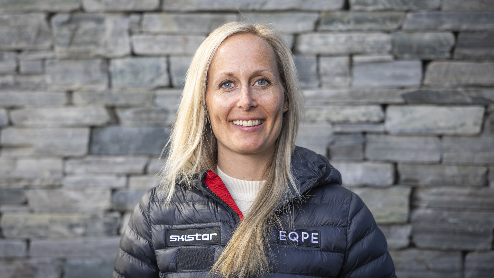 Kvinna i svart jacka med texten "Skistar" och "EQPE", står framför en stenvägg