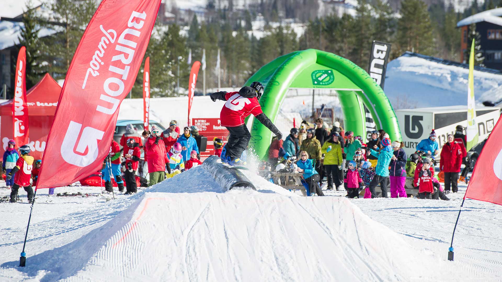 Snowboardtävling för barn med åskådare i en vintersportanläggning, omgiven av färgglada fanor och snötäckta landskap