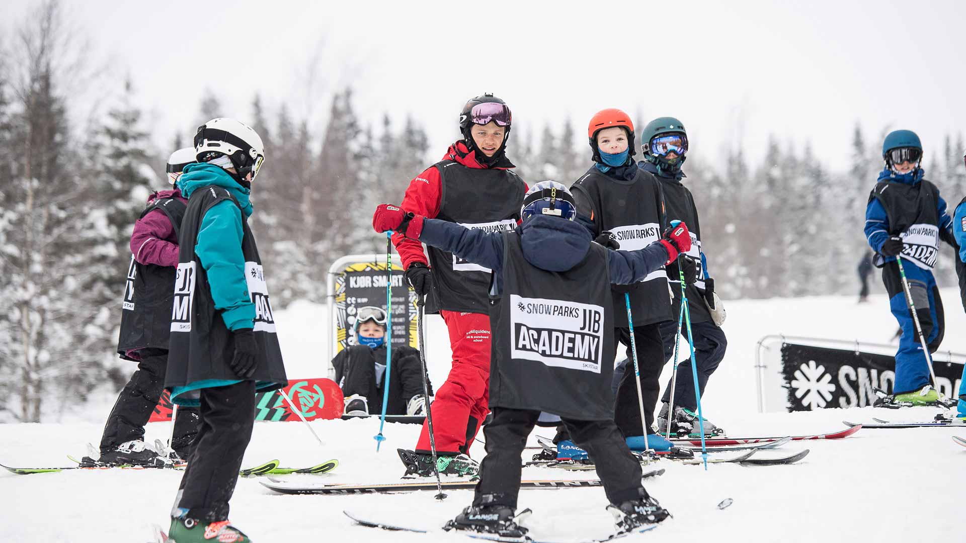Skidinstruktör i röd dräkt undervisar en grupp barn i skidåkning på en snöig backe under SkiStar Jib Academy