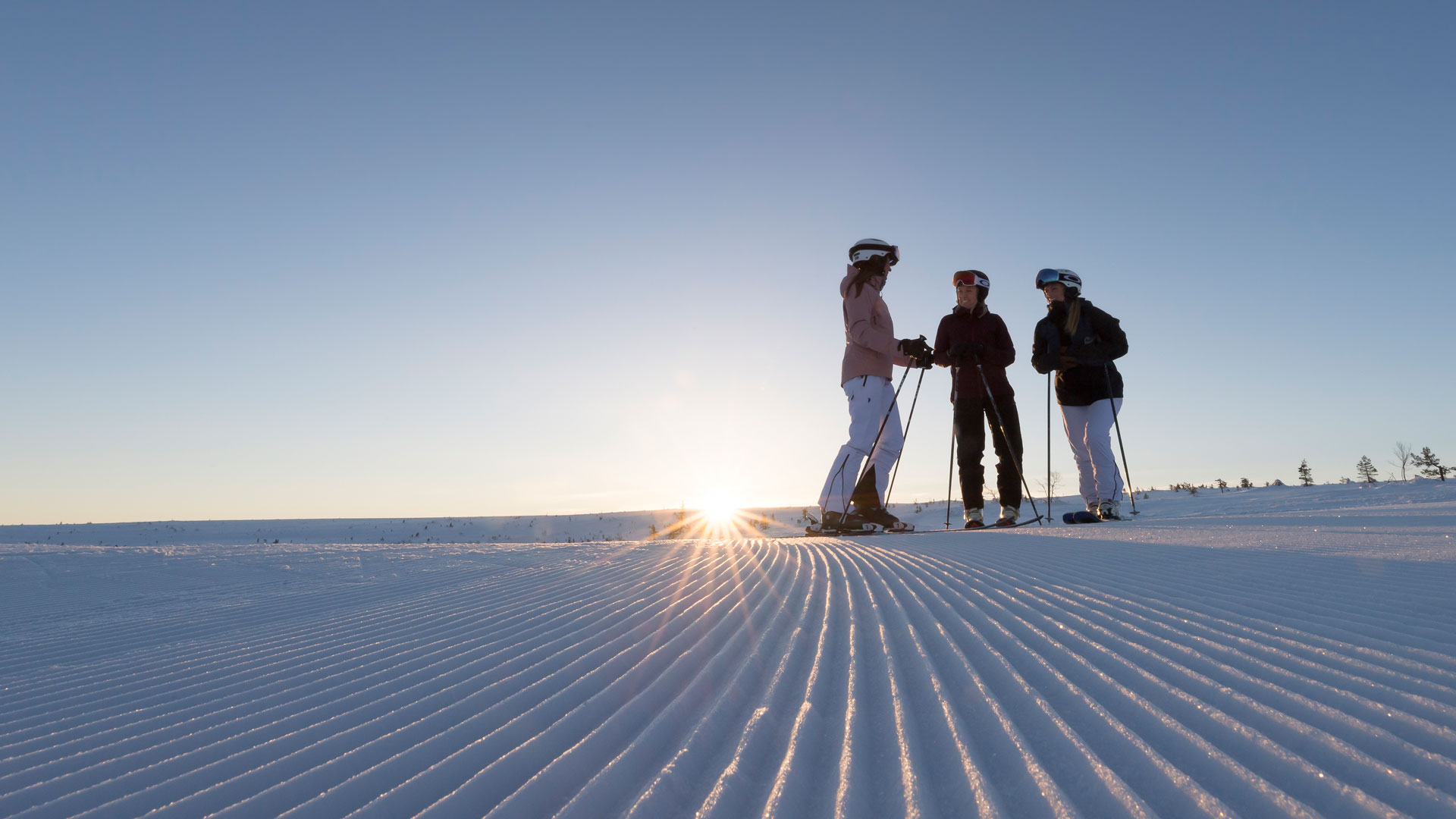 Tre skidåkare samtalar på en preparerad skidbacke vid solnedgången