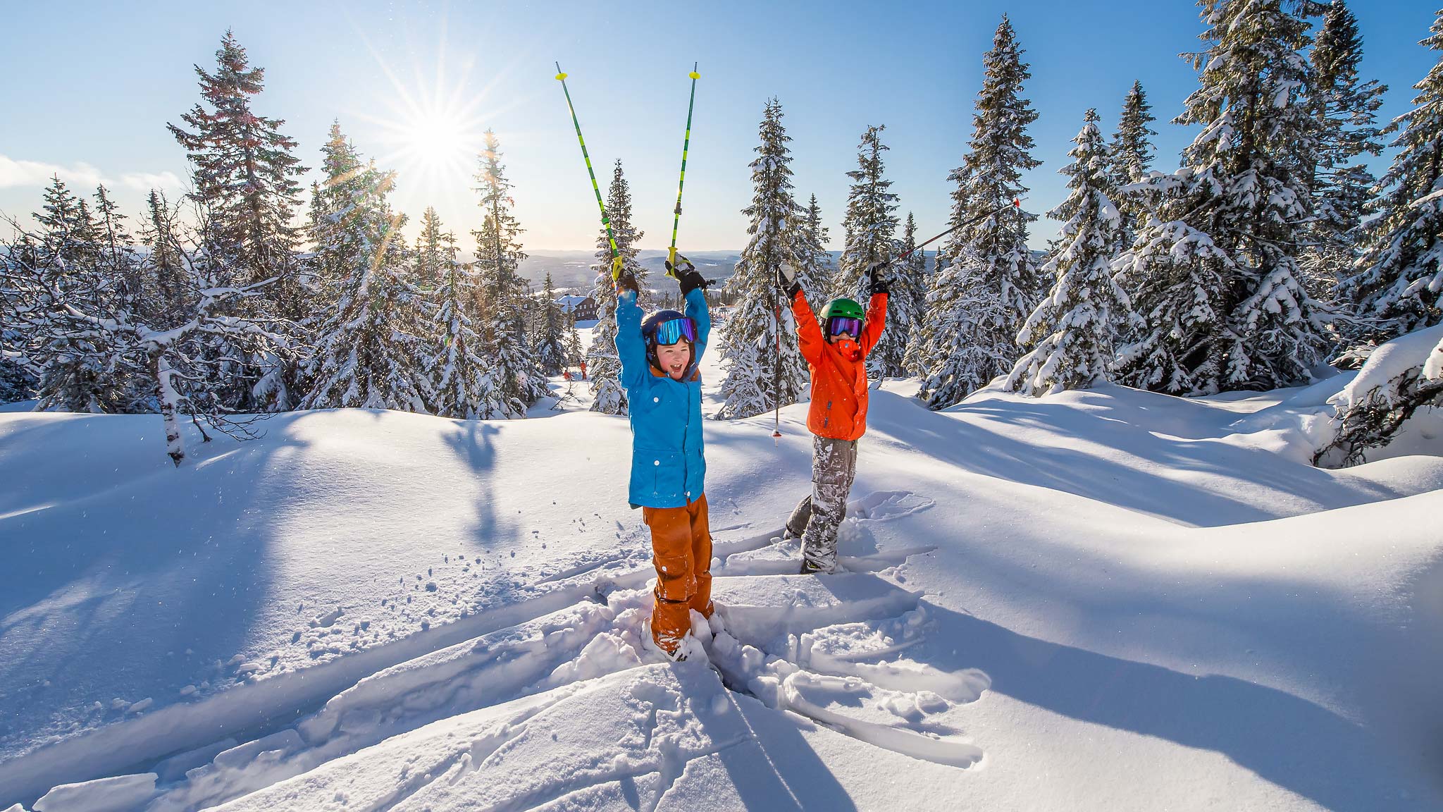 Två glada barn som åker skidor i nysnö, omgivet av snötäckta träd och soligt vinterlandskap