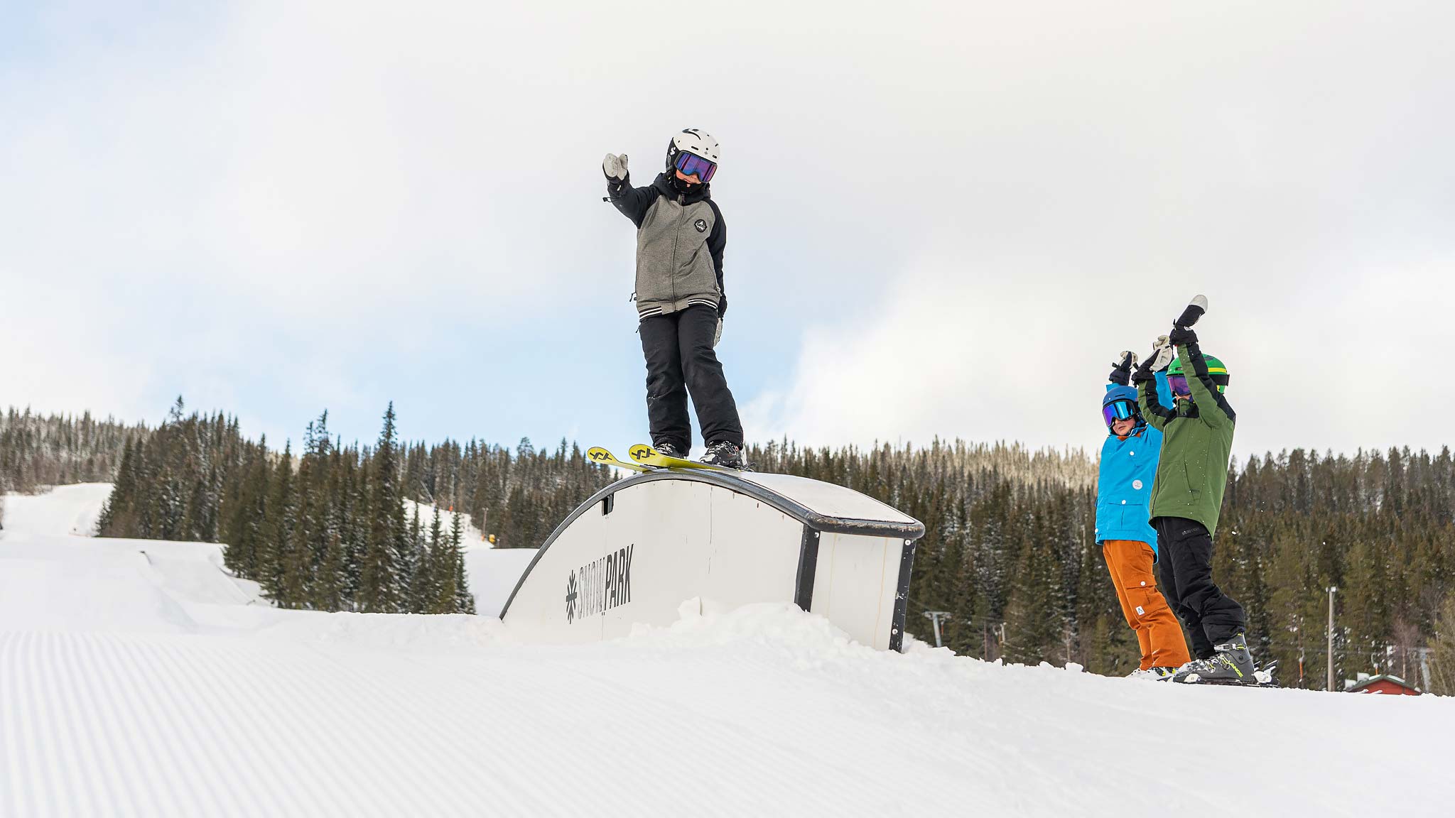 Person som åker snowboard över en snöräls i en vinterpark, med två personer som jublar i bakgrunden. Skogsklädda berg syns i fjärran under en molnig himmel
