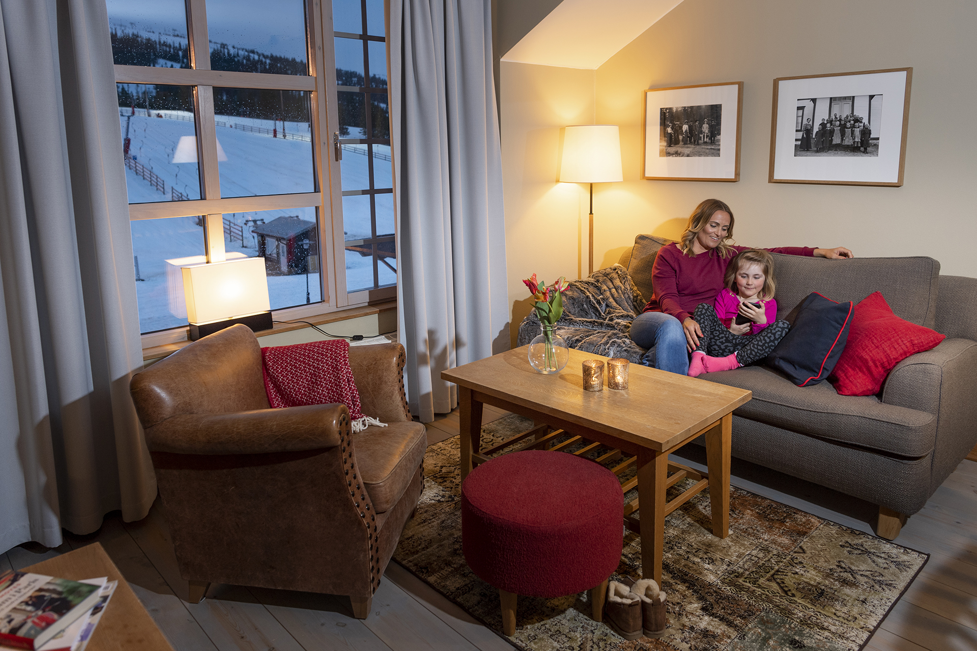 Personer i en mysig stuga med utsikt över snöiga berg, sitter i soffan och använder en smartphone. Inredningen består av en fåtölj, soffbord och varma textilier