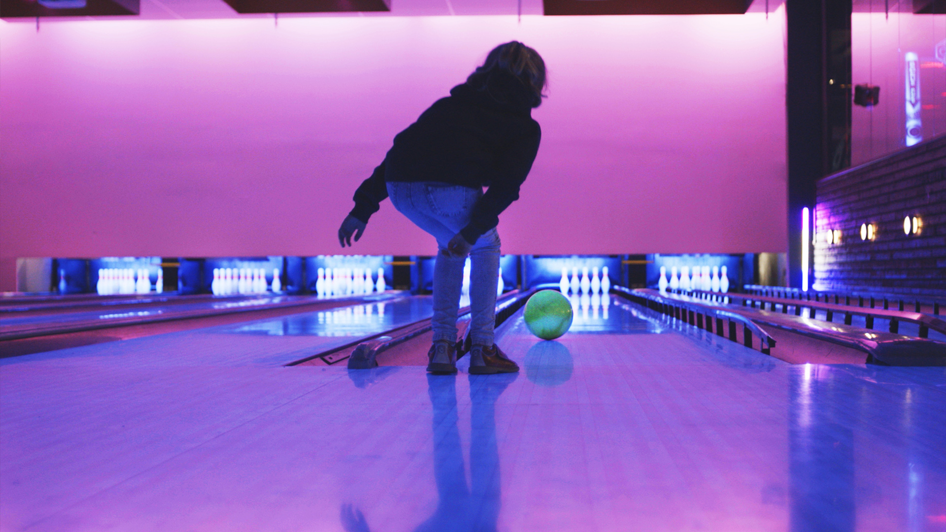 Person som spelar bowling i neonupplyst bowlinghall, rullar klot mot käglor på banan