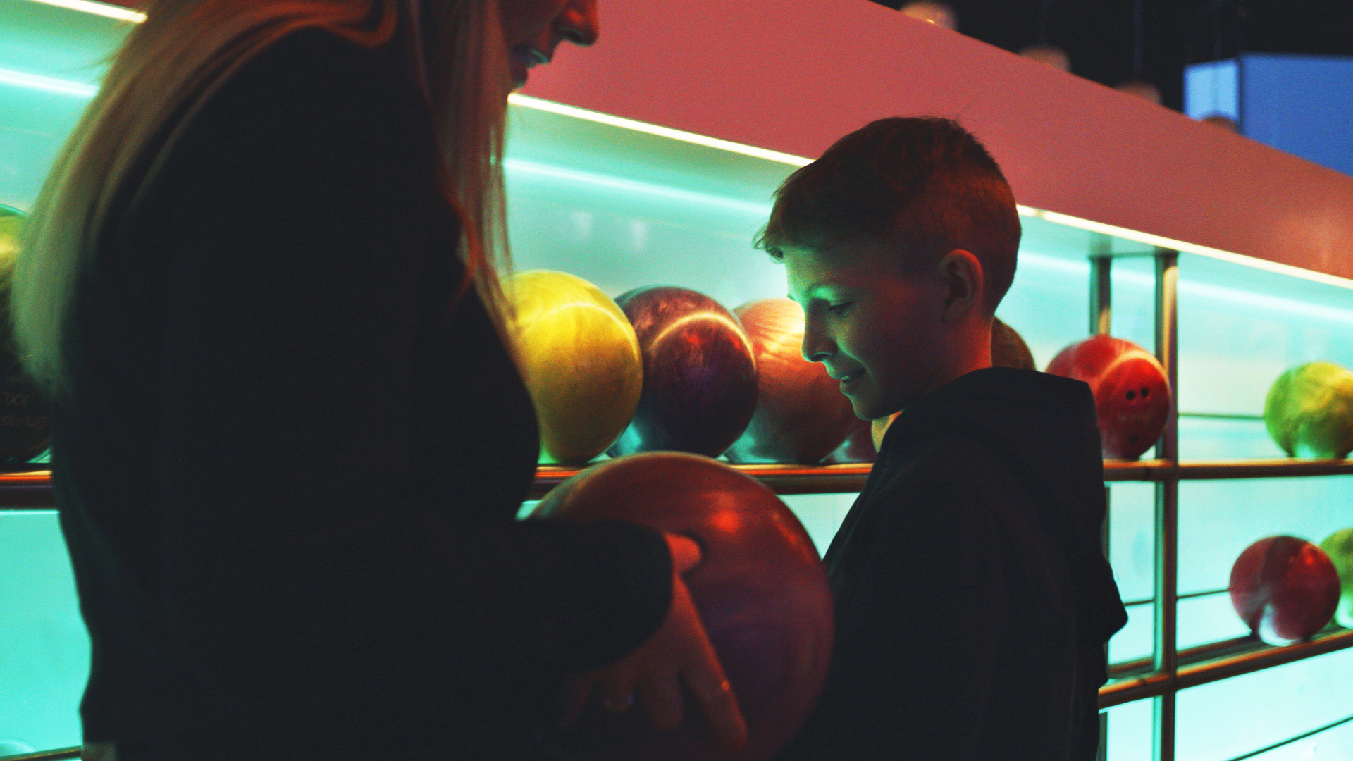 En kvinna och ett barn vid en bowlinghall plockar bowlingklot från en lysande hylla