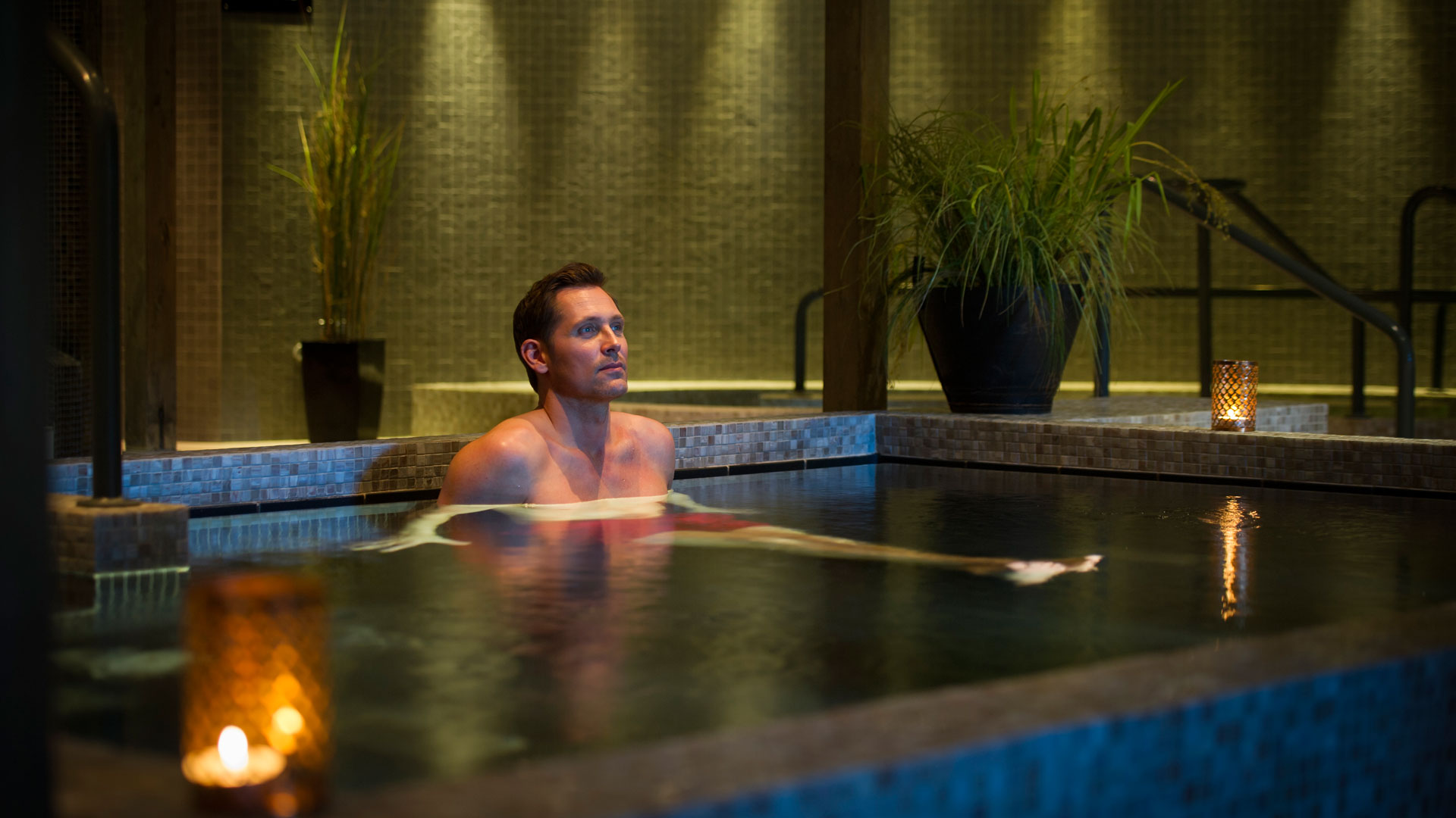 Man njuter av avkoppling i ett spa med dämpad belysning, omgiven av gröna växter och levande ljus