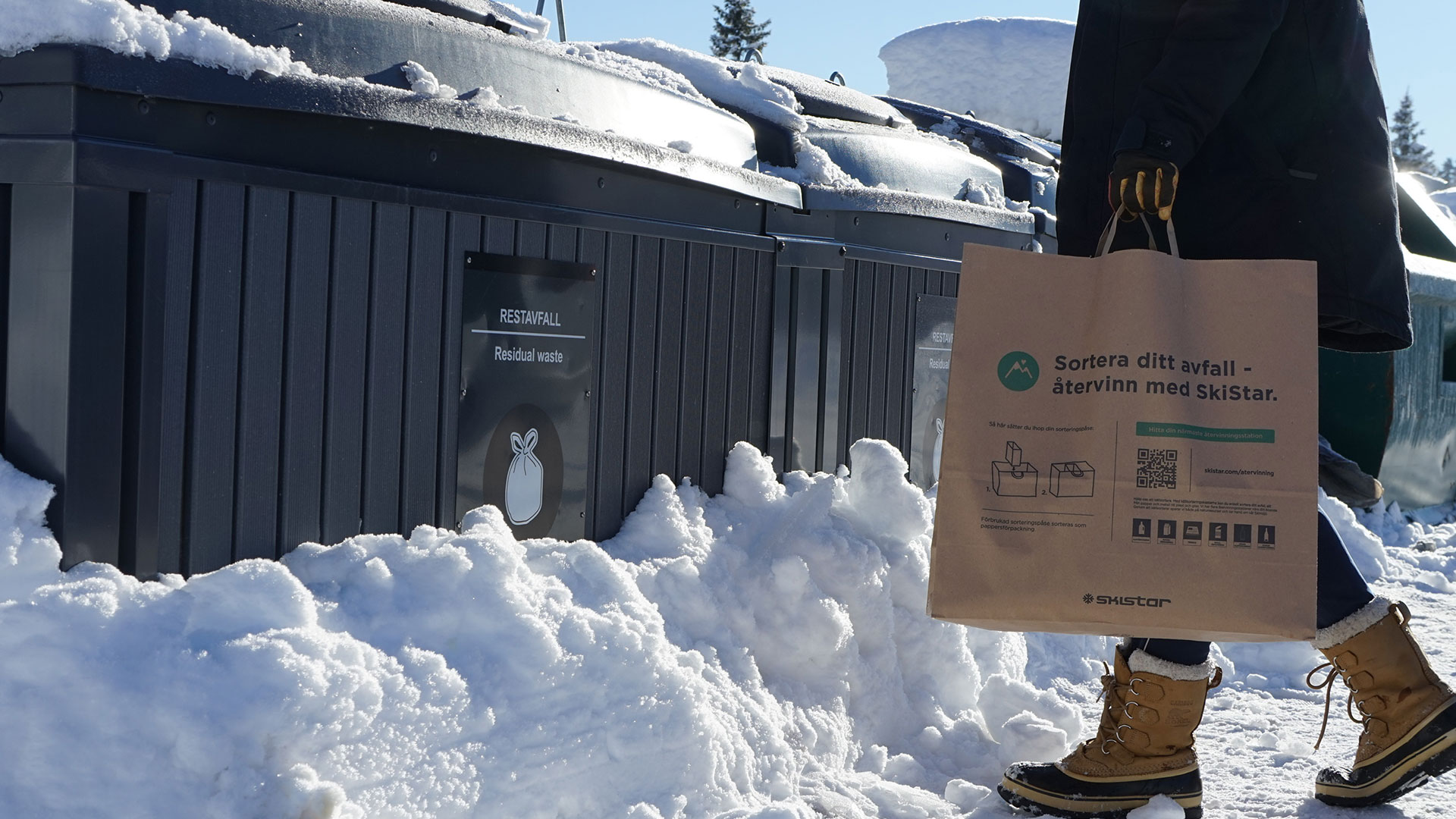 Person bär papperspåse för återvinning framför sopcontainer i snötäckt vintrig miljö