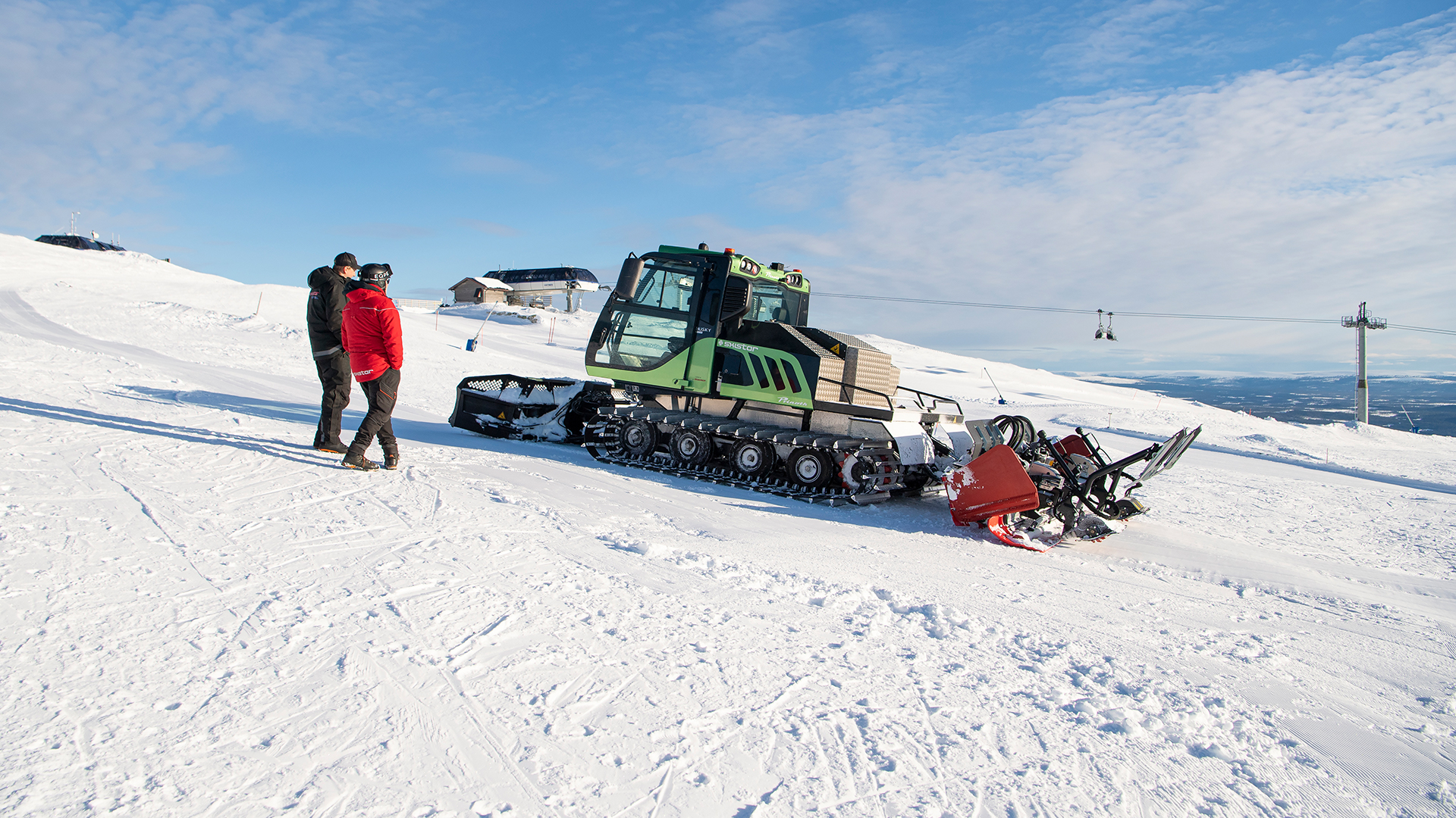 Grön elpistmaskin på snötäckt fjäll, två personer i samtal bredvid den, under klar blå himmel