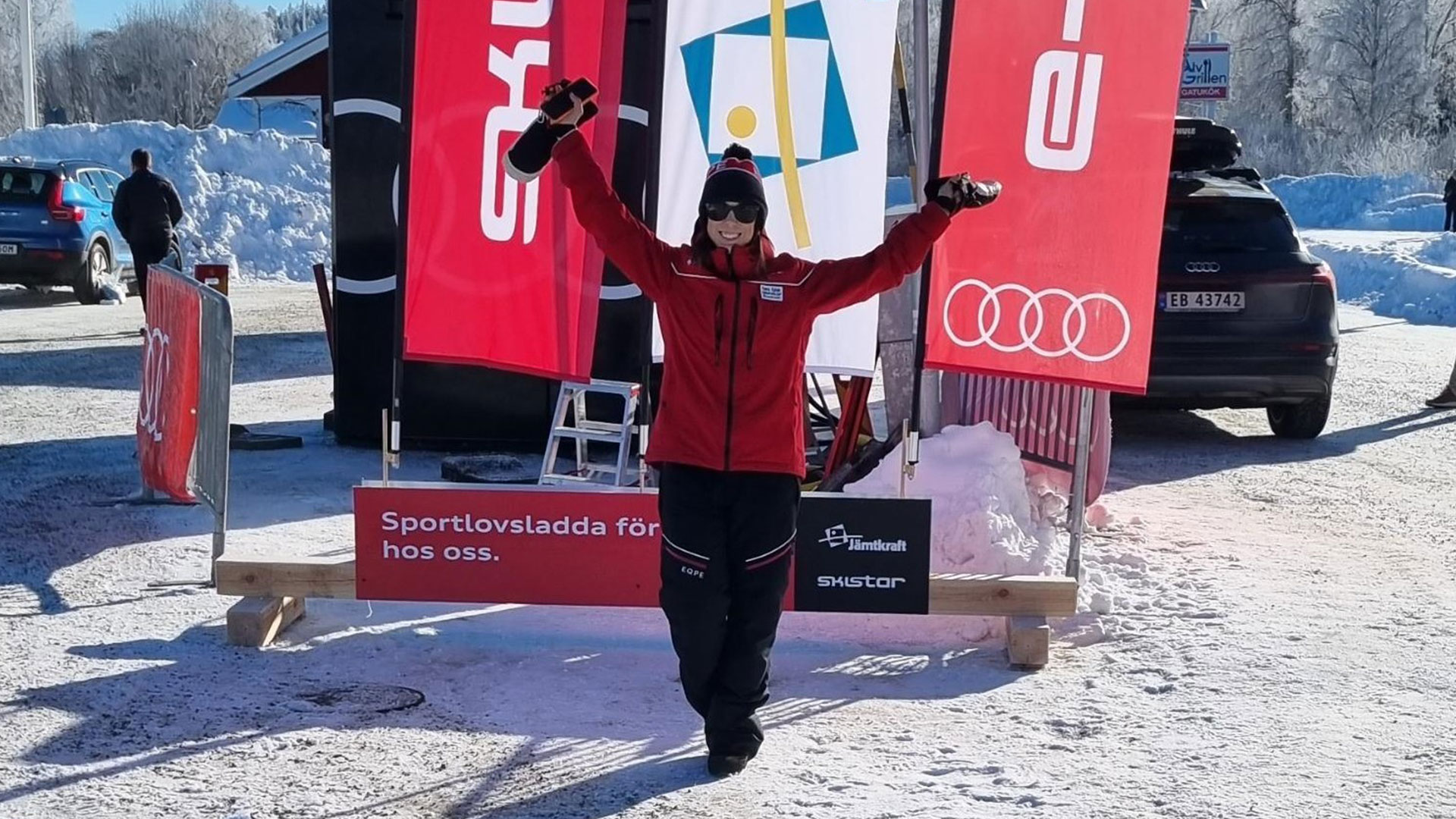 Person i röd jacka jublar framför Audi- och Skistar-skyltar på ett snötäckt sportlovsevenemang