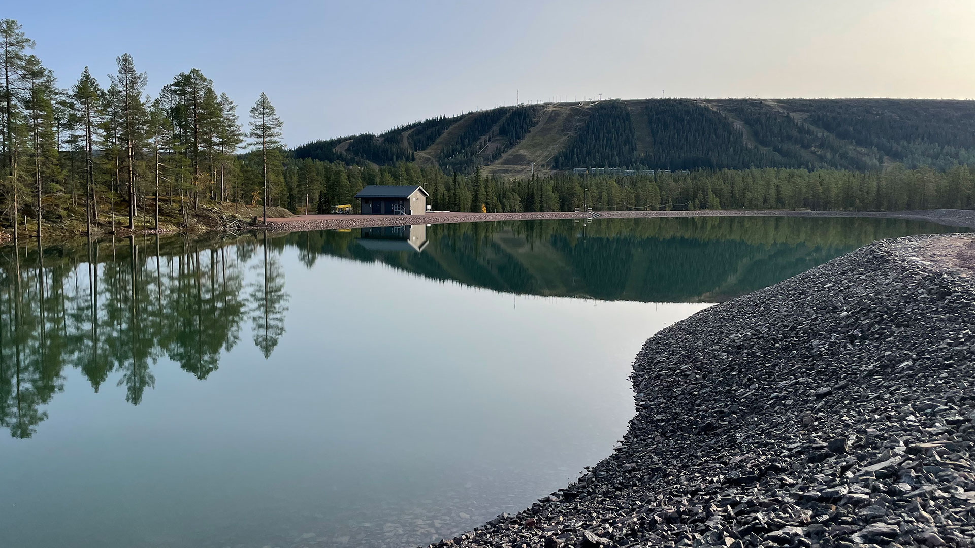 Sjön med klart vatten och skog i bakgrunden, med en liten stuga vid strandkanten under en klar himmel