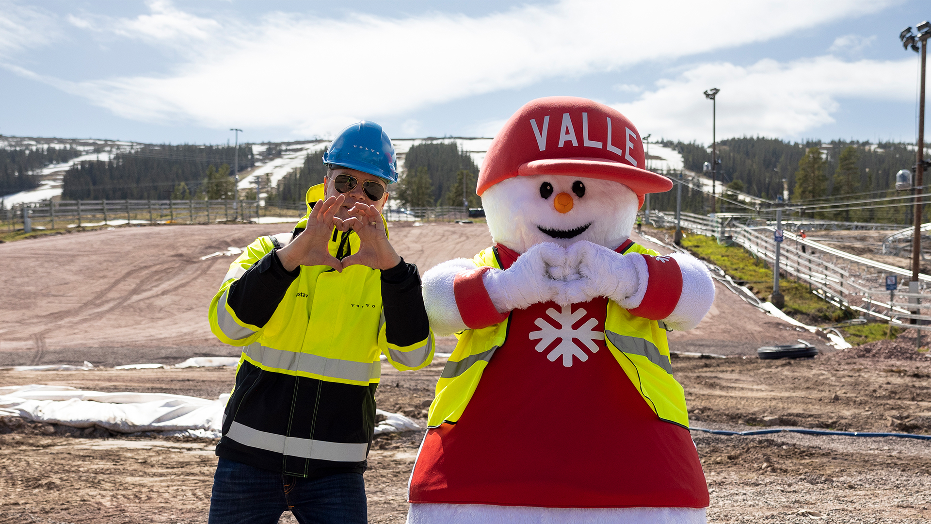 Person i reflexjacka bredvid en stor snögubbe-maskot med hjälm och väst, båda gör hjärttecken med händerna på en skidort under klar himmel