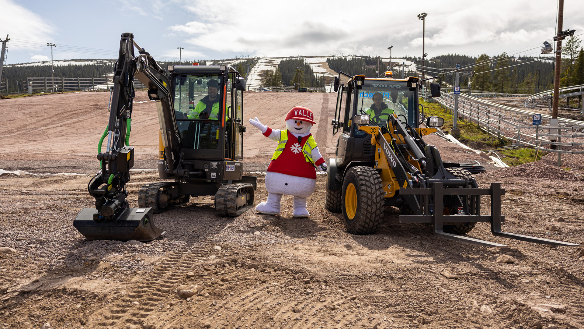 Maskot i skidområde med två anläggningsmaskiner, Volvo utrustning omgiven av skogslandskap