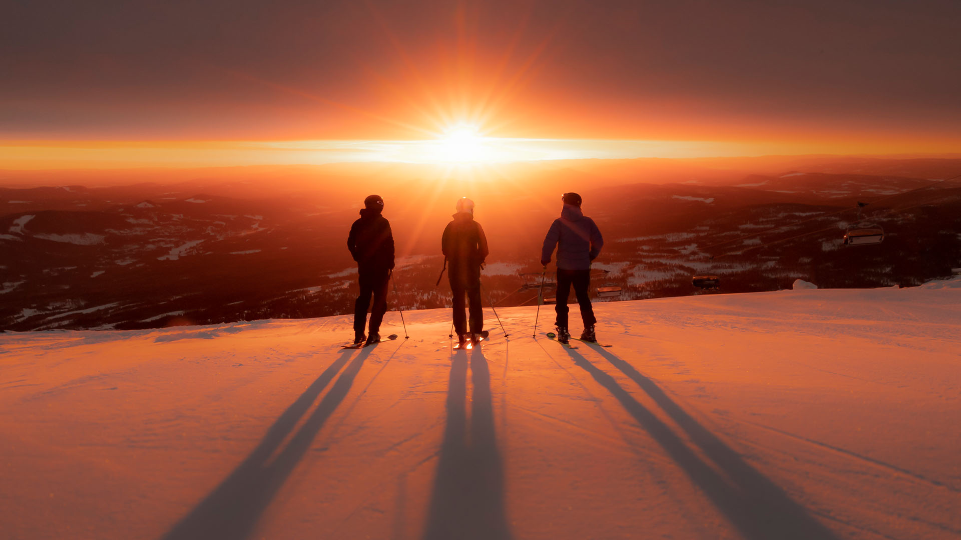 Tre personer står på en snötäckt bergstopp vid solnedgången, redo att åka skidor