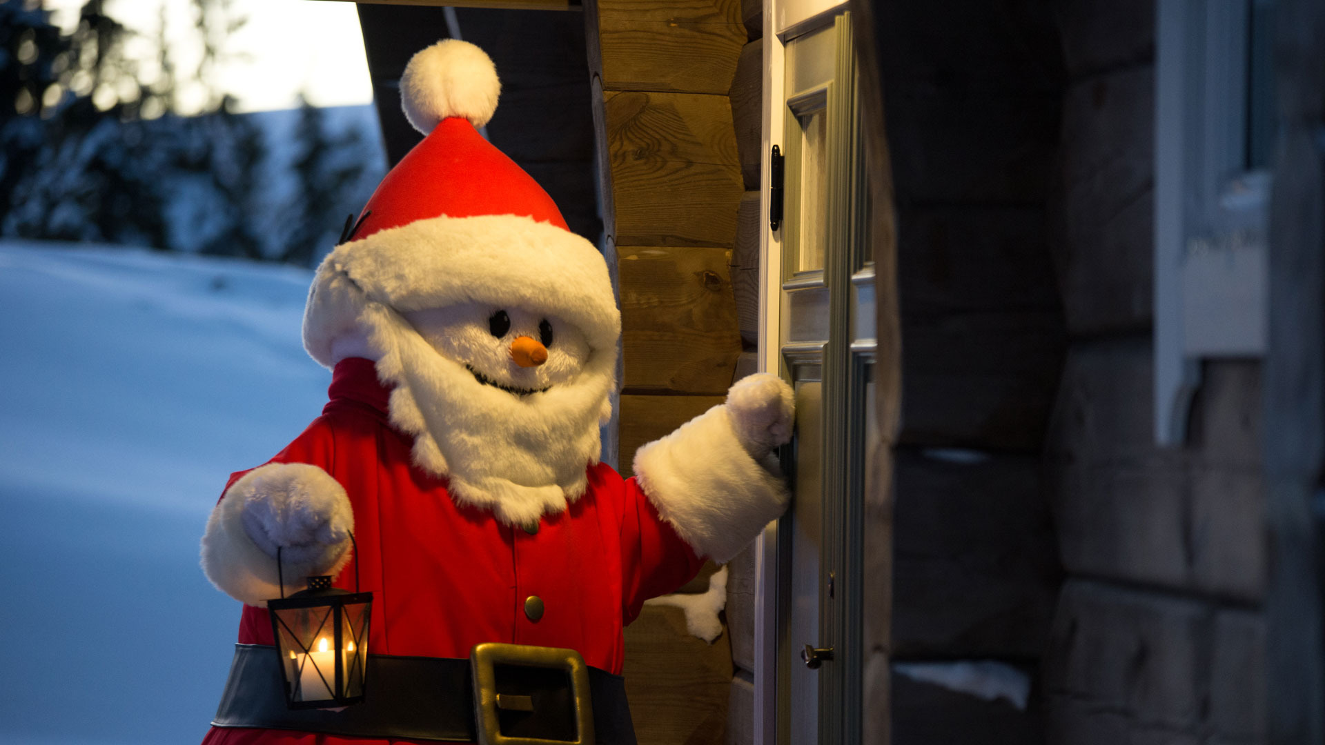 Tomte i röd dräkt med lykta knackar på dörren till en stuga, omgiven av snöig vinterlandskap