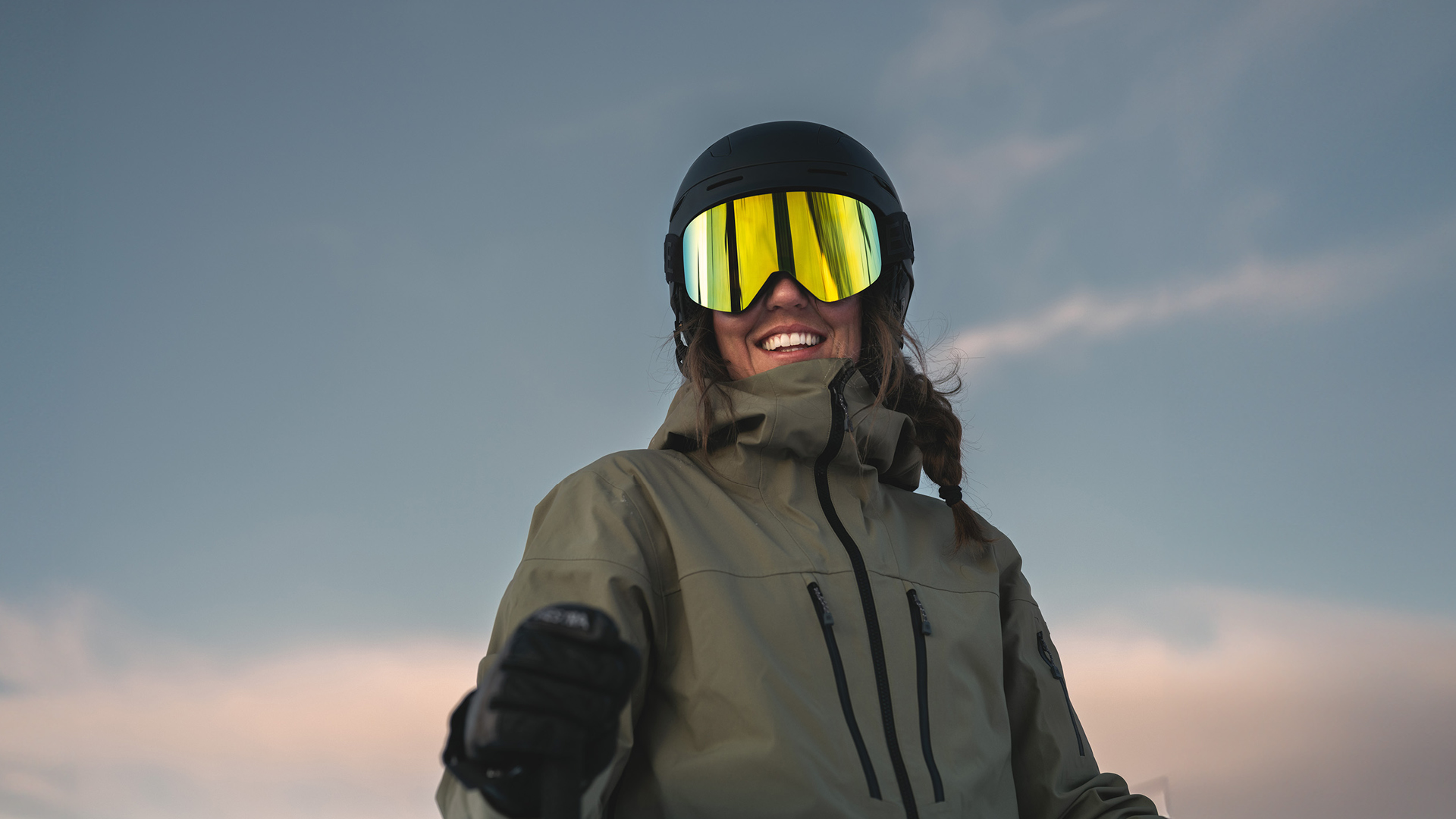 Person med skidglasögon och hjälm som åker skidor, klädd i vinterjacka, mot en klar himmel