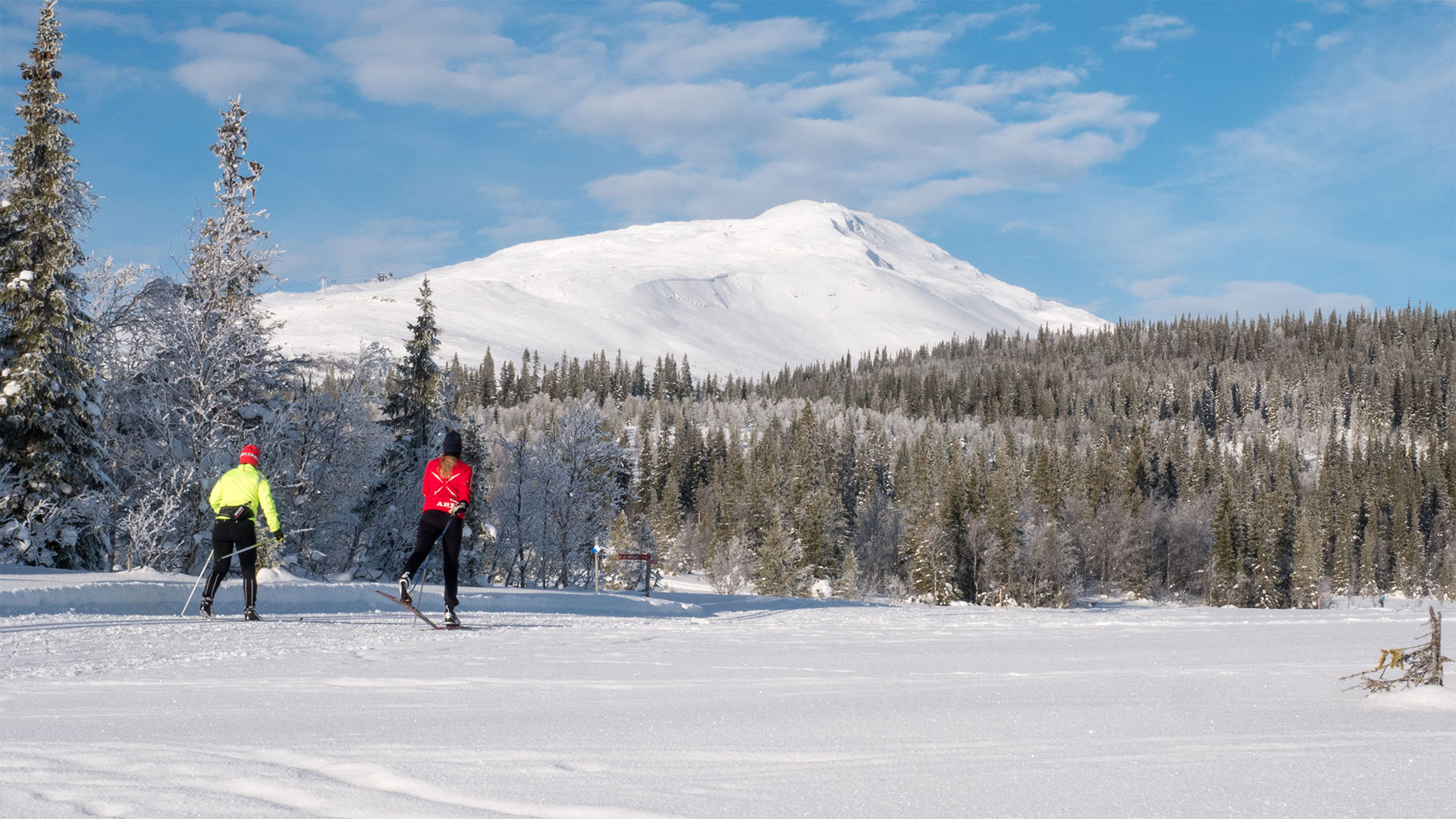 Två längdskidåkare på vinterstig med snöbeklädda berg i bakgrunden, omgivna av skog