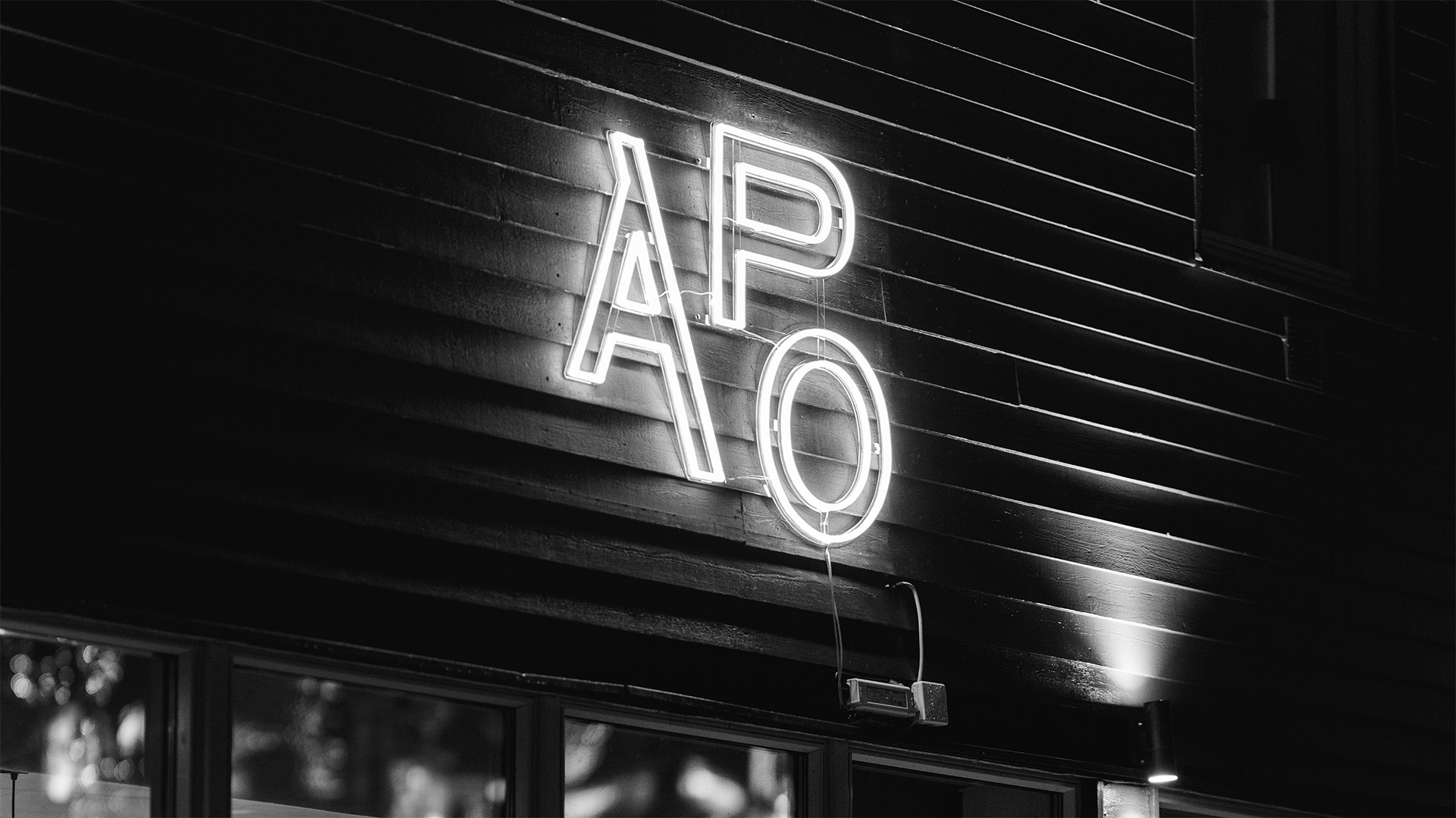 Neonskylt med bokstäverna "APO" på en mörk byggnadsvägg i svartvitt