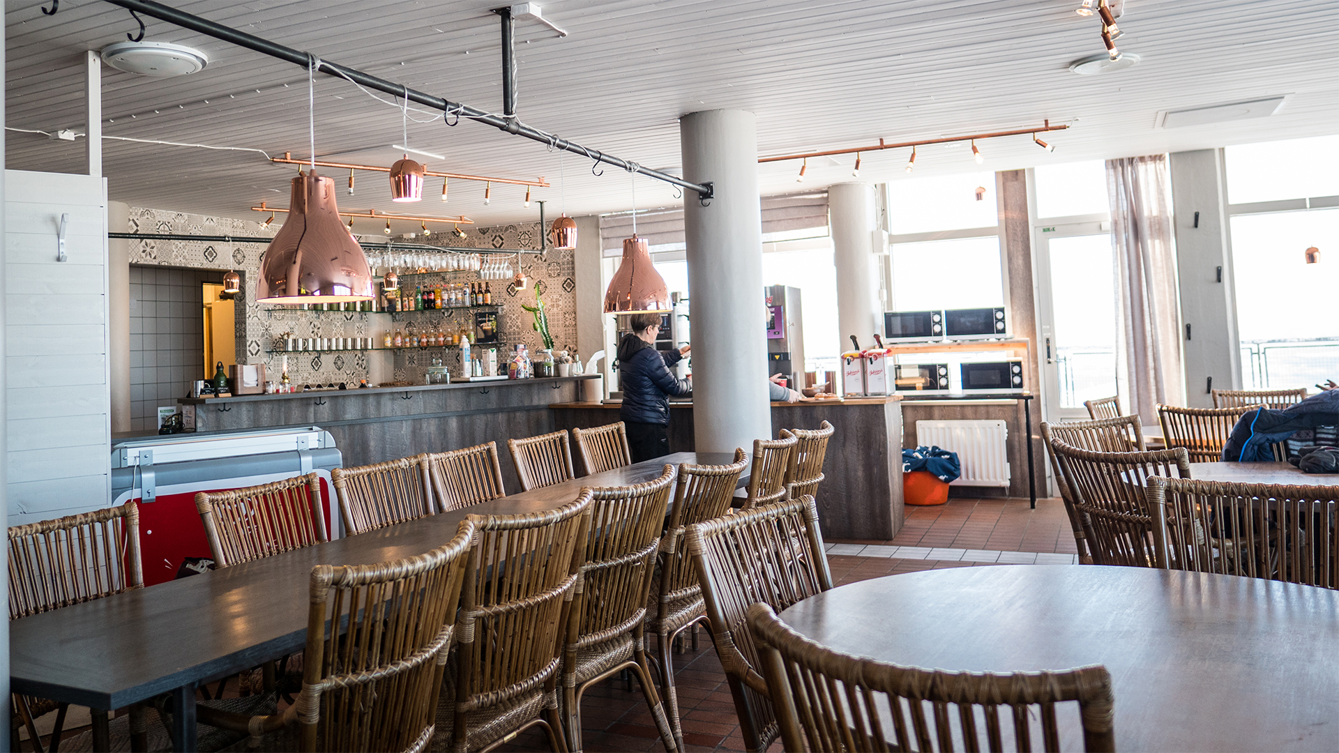 Restauranginteriör med korgstolar, träbord och en välfylld bar i bakgrunden. Ljusa lampor hänger från taket
