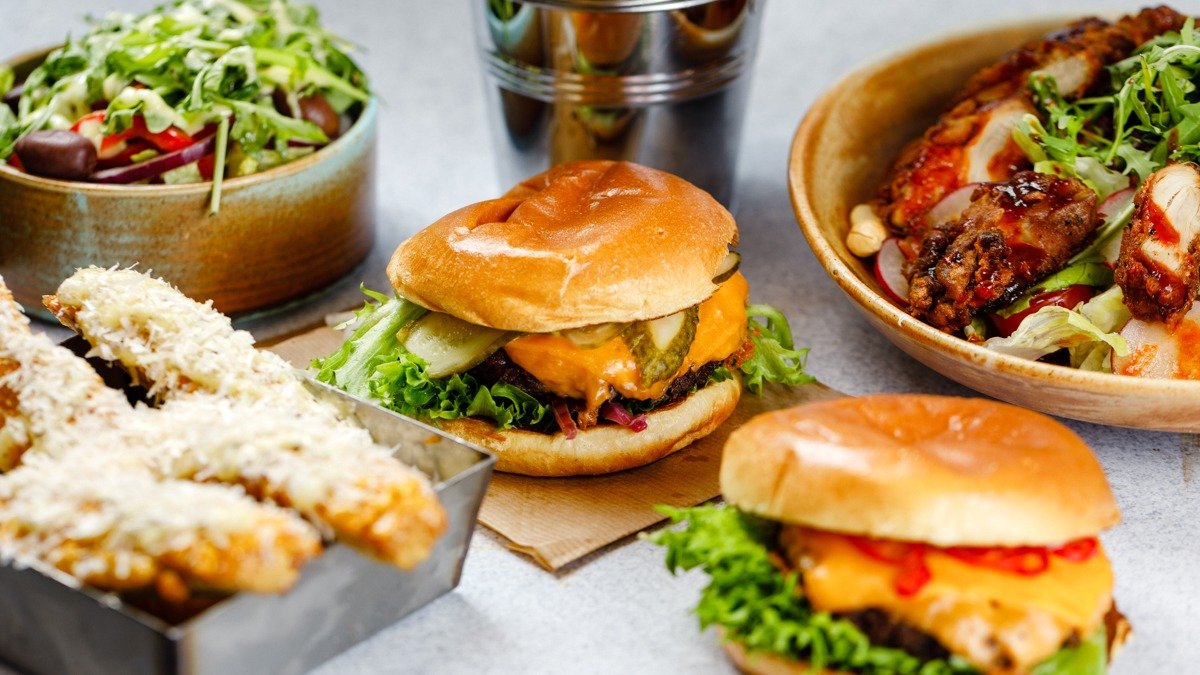 Två hamburgare med ost, pickles och sallad, sötpotatisfries med parmesan, och en fräsch sallad med grönsaker och grillad kyckling