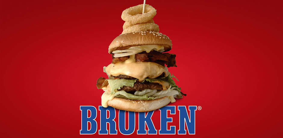 Stor cheeseburgare med dubbla köttpuckar, bacon, sallad, ost, och lök på röd bakgrund, märkt med "Broken" längst ner