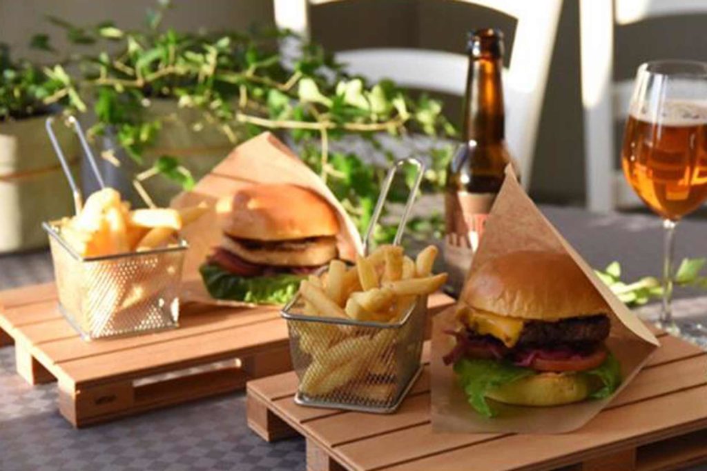 Hamburgare med pommes frites på träbrickor, flaska öl och ölglas i bakgrunden, omgiven av gröna växter