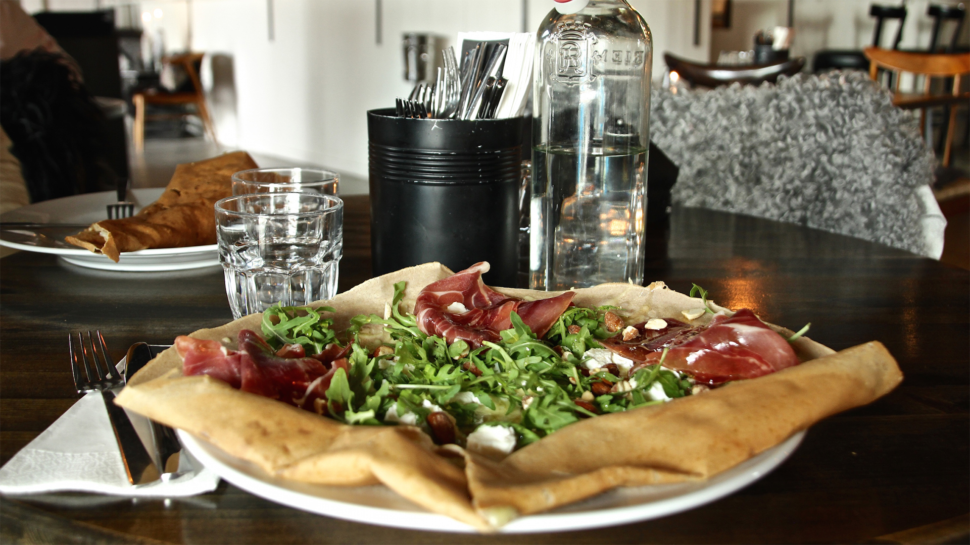 Tallrik med galette toppad med ruccola och parmaskinka på rustik restaurang, med vattenflaska och glas i bakgrunden