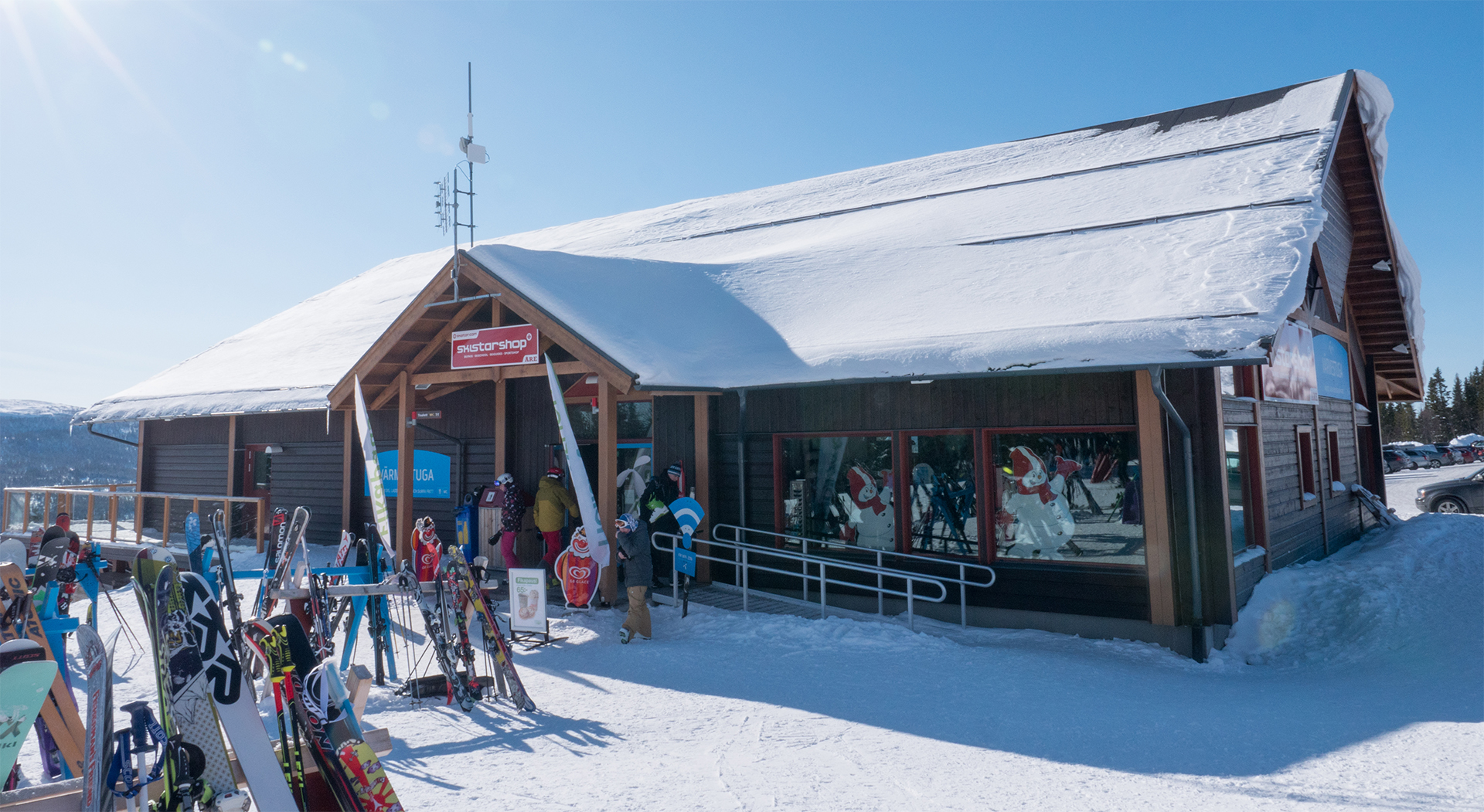 Holiday Shop Rödkullen, skidbutik och stuga i vintrigt landskap. Skidor står utanför byggnaden, som har ett snötäckt tak under klarblå himmel