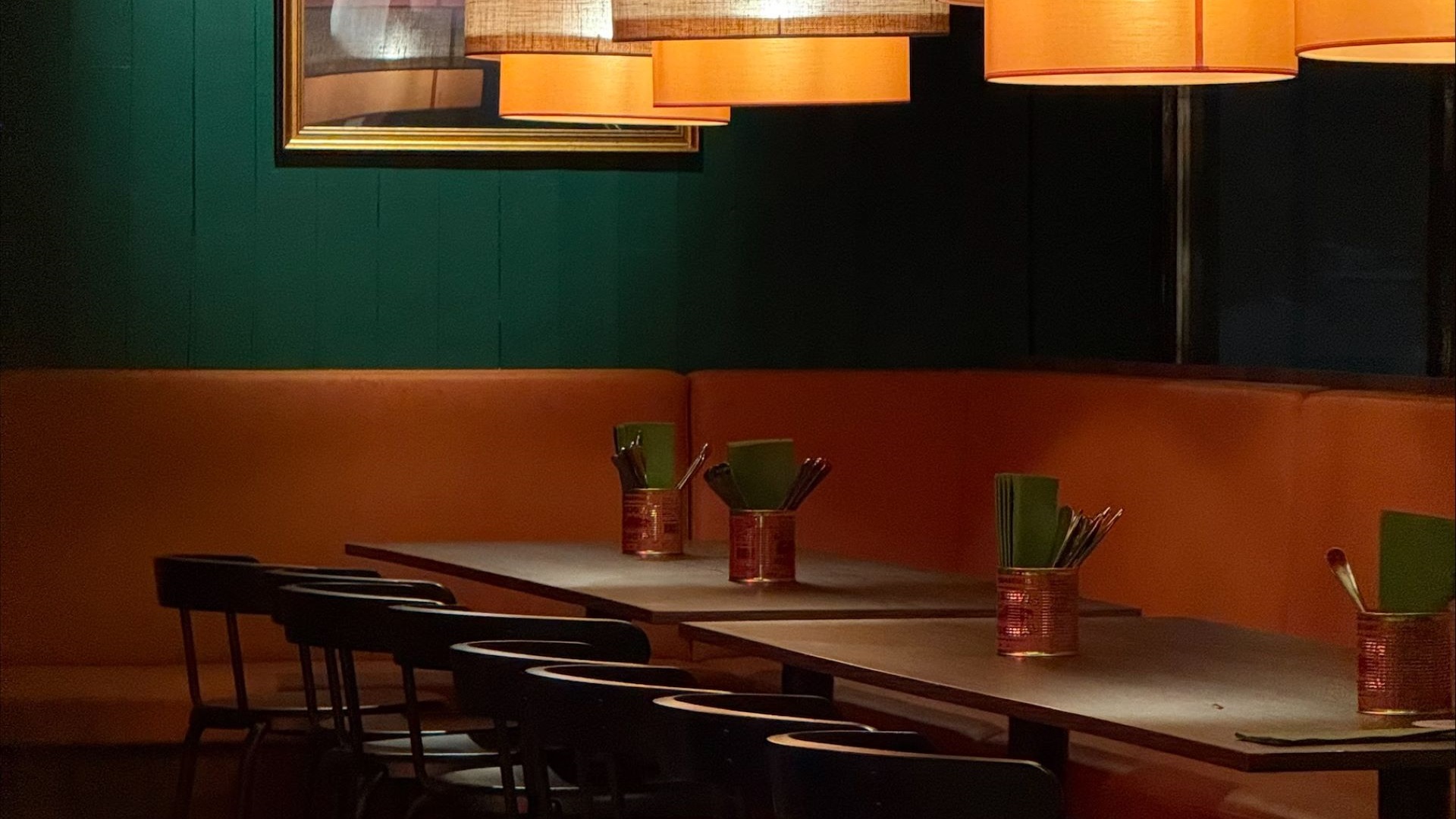 Restauranginteriör med mörkgrönt väggpanel och orange stoppade sittbänkar. Bord med stolar och röd metallhållare med menyer. Ovanför hänger runda lampor med varma ljus