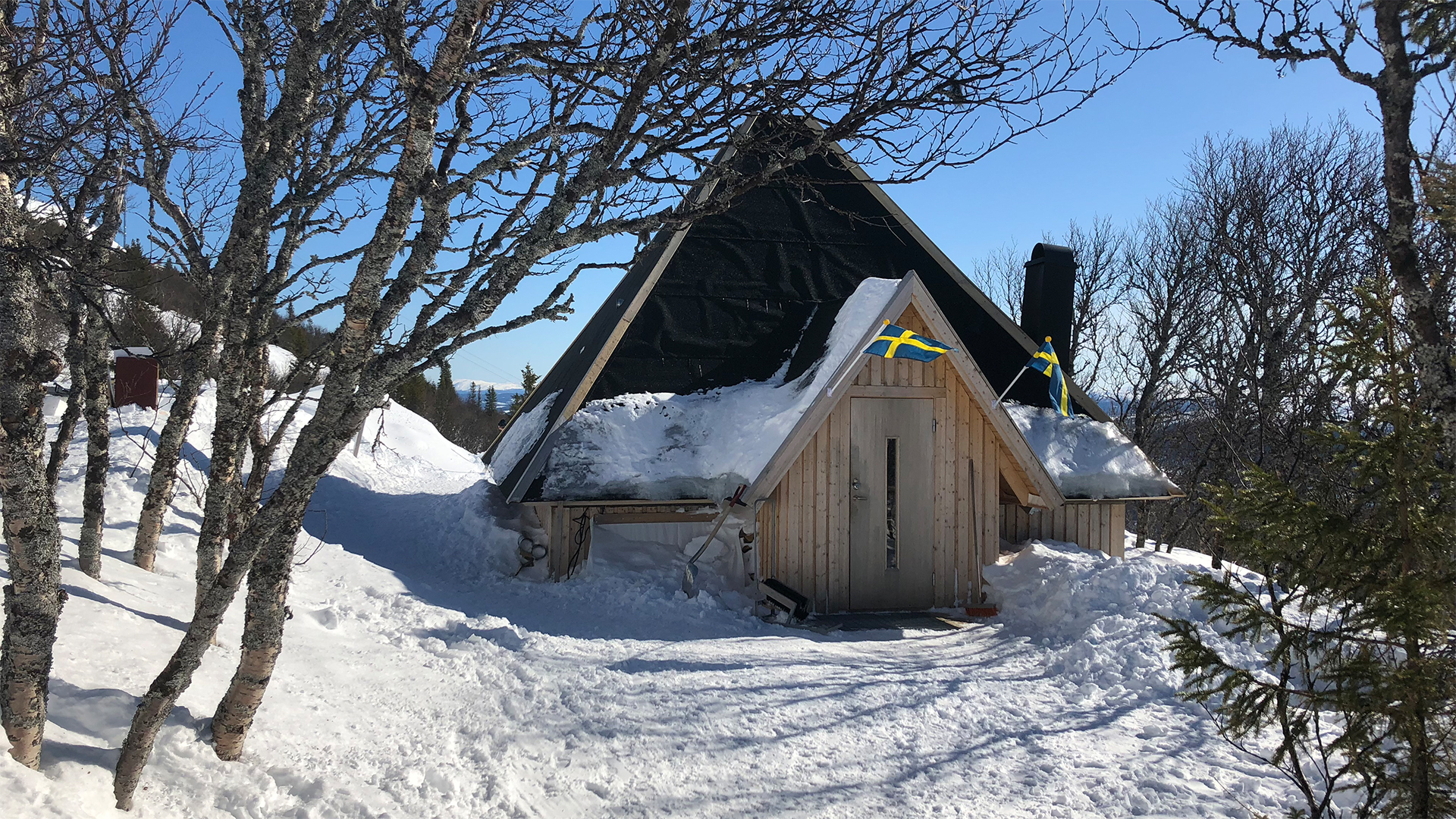 Svensk fjällstuga täckt av snö en solig vinterdag med svenska flaggor, omgiven av snötäckta träd och blå himmel