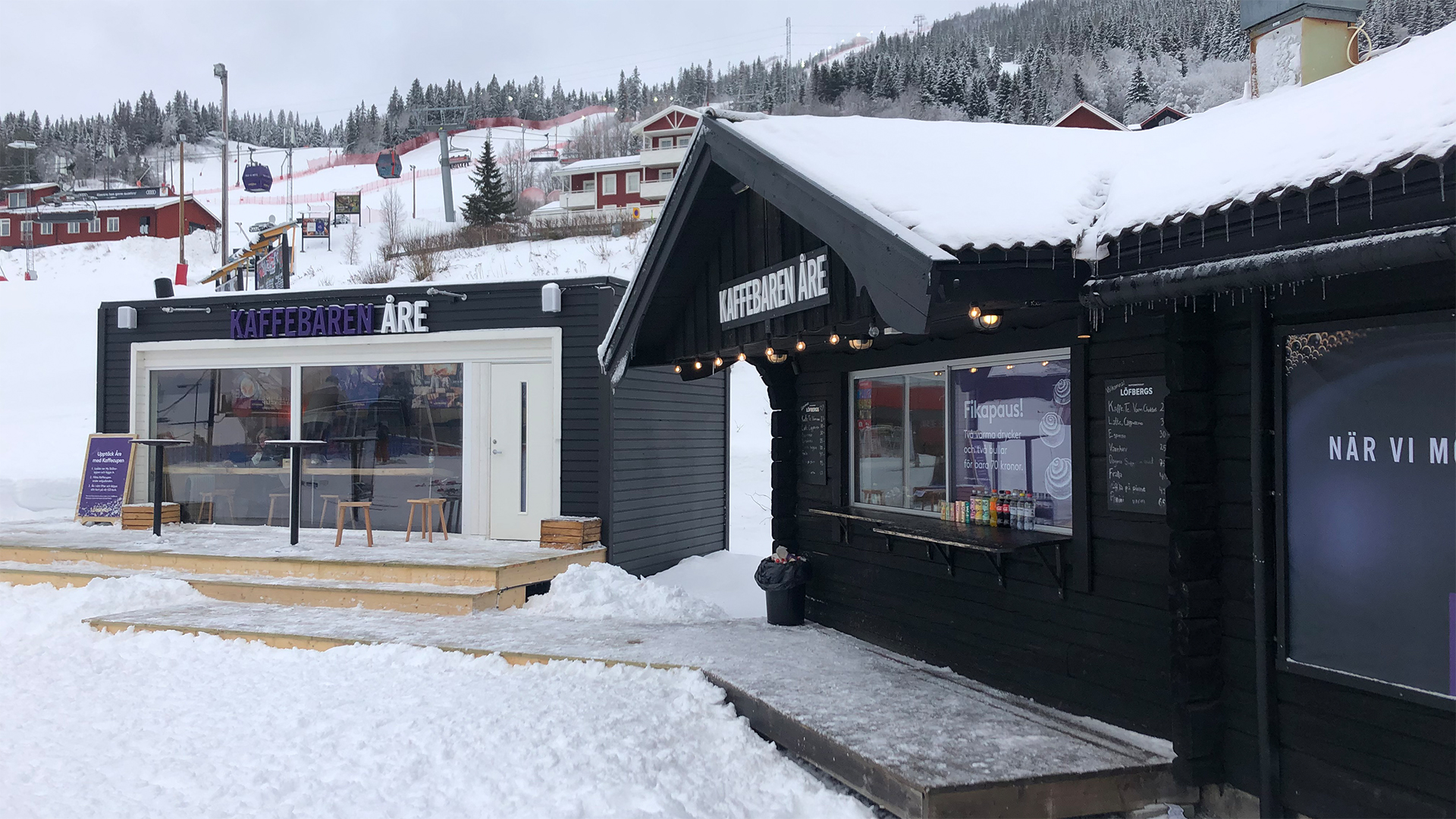 Kaffebaren Åre i snölandskap, med två timrade byggnader och skidbacke i bakgrunden i Åre, Sverige