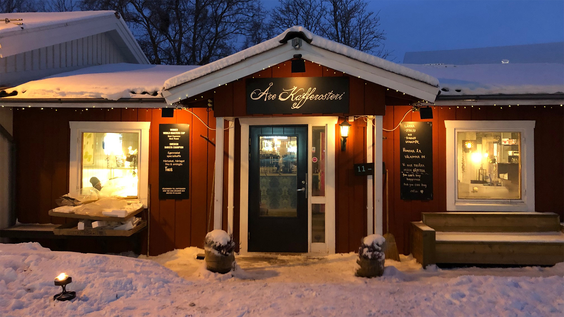 Snötäckt byggnad av Åre Kafferosteri belyst på kvällen med skyltar och fönster, omgiven av vinterlandskap