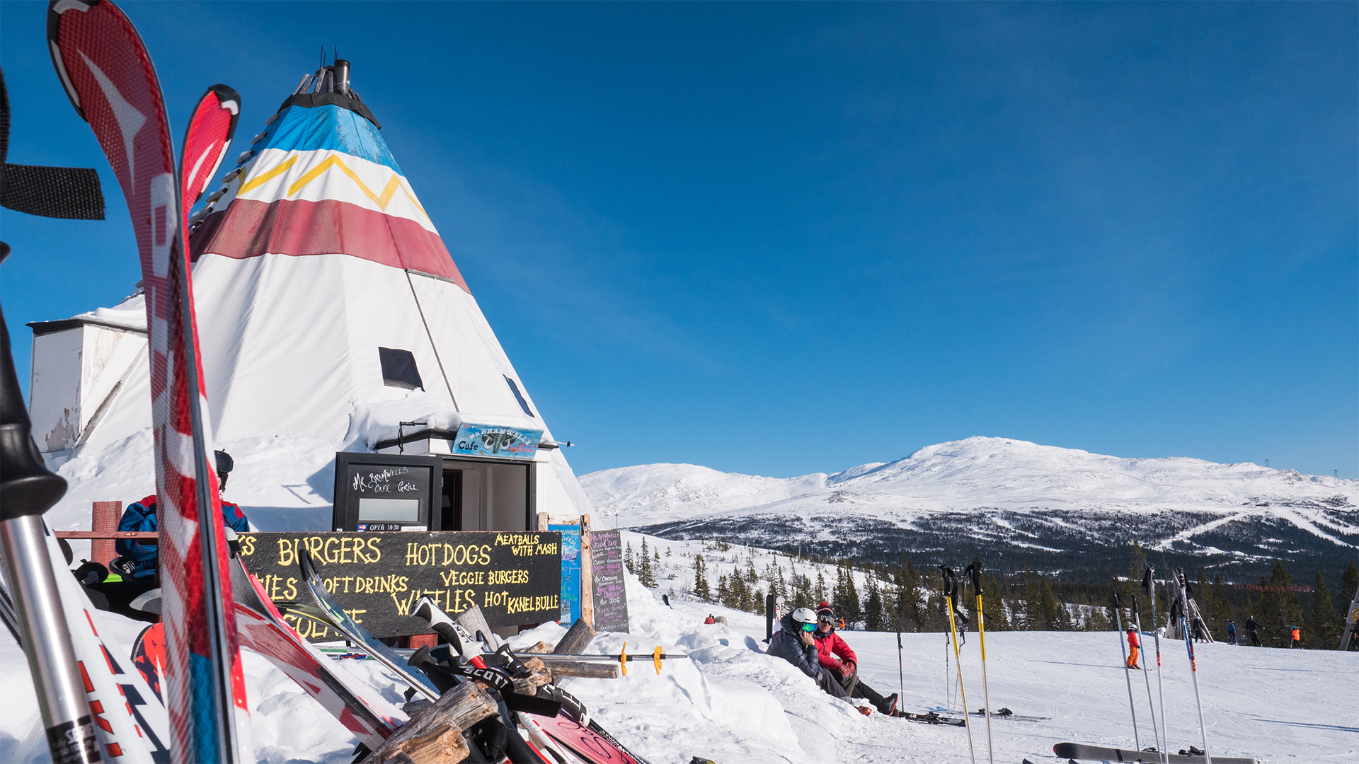 Skidåkare vid snötäckta fjäll med kafétält, Åre, Sverige. Skidor står lutade mot tältet under klarblå himmel, perfekt för vinteraktiviteter